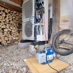 Instalacja zewnętrznej jednostki grzewczej z otwartą obudową, podłączona do pompy próżniowej VALUE na drewnianej płycie, w tle stos drewna i rower.