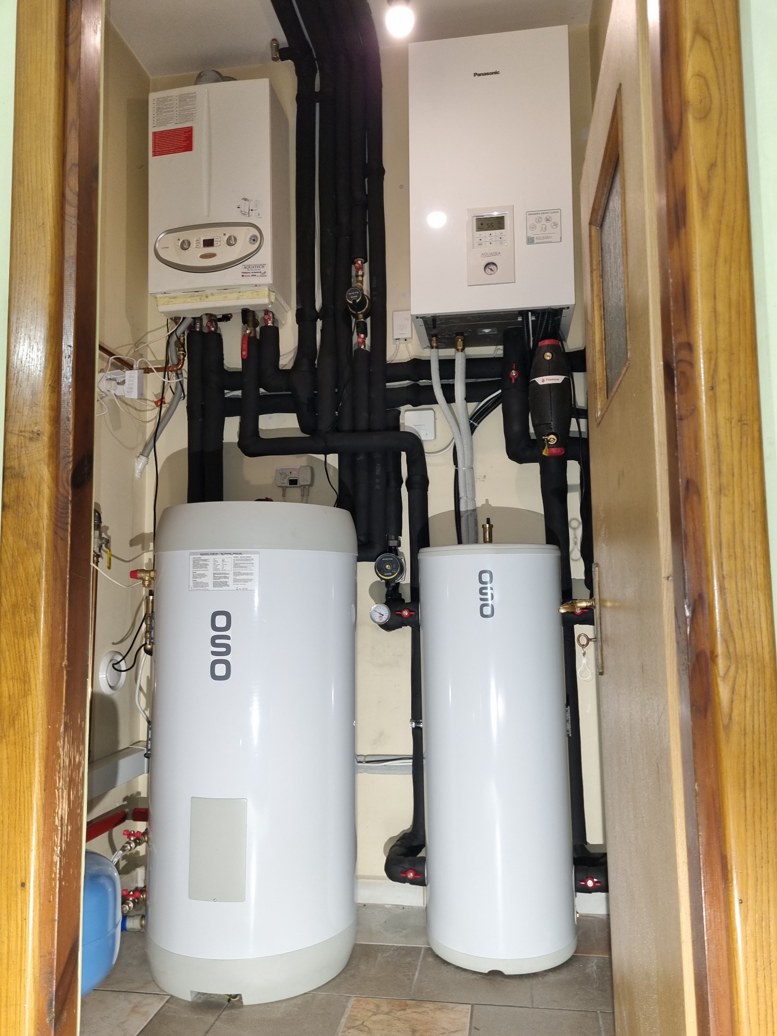 Kompaktowa kotłownia z dwoma białymi zasobnikami ciepłej wody OSO, dwoma wiszącymi kotłami Aquatech i Panasonic, oraz rozbudowaną instalacją rur w izolacji, umieszczona w małym pomieszczeniu...