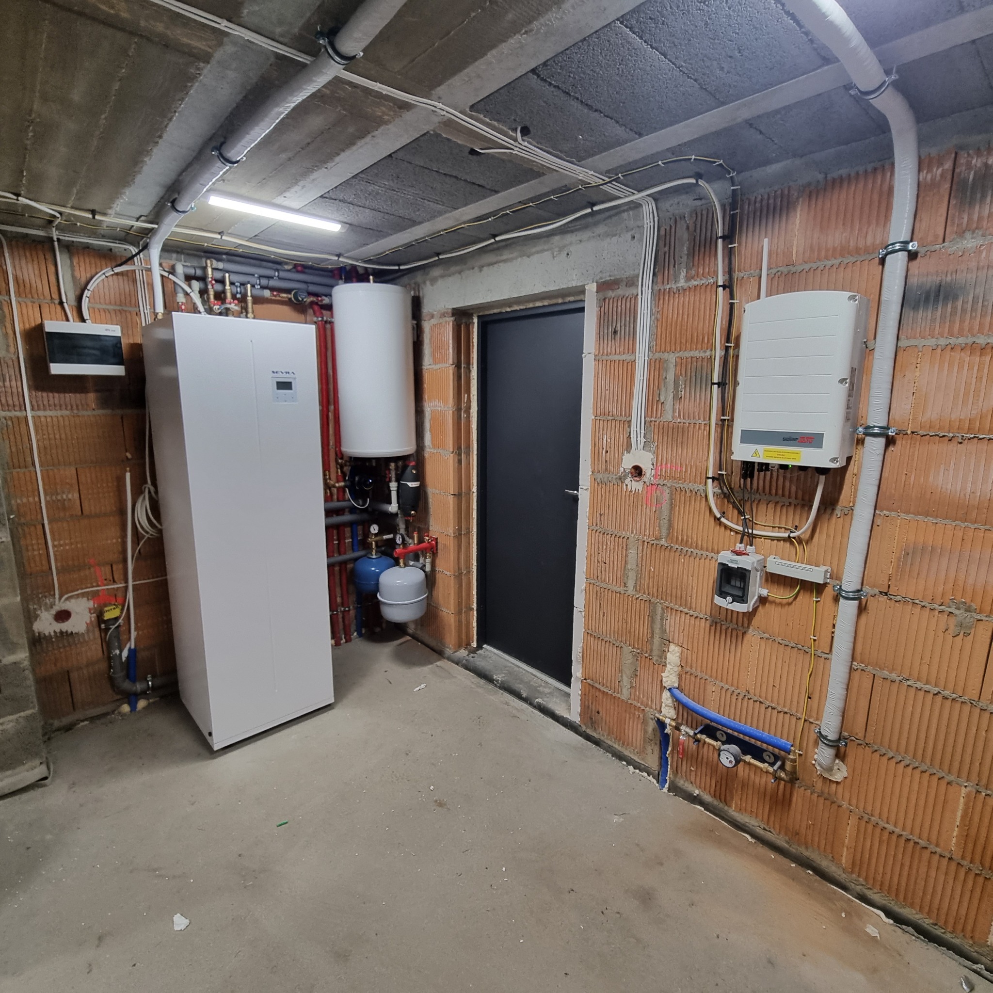 Kotłownia w surowym stanie: biała jednostka grzewcza z zbiornikiem buforowym, falownik fotowoltaiczny na ceglanej ścianie, instalacja elektryczna i hydrauliczna w szarym pomieszczeniu z betonowym...