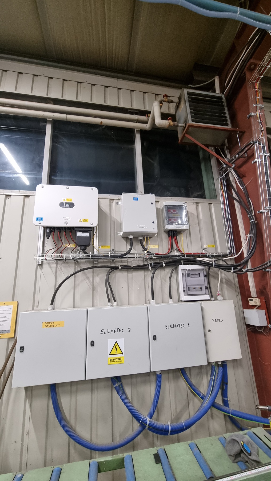 Instalacja elektryczna z rozdzielnicami i przewodami na ścianie w hali, widoczny inwerter, zabezpieczenia i rury ochronne. Tabliczki ostrzegawcze na obudowach. U góry okno i grzejnik przemysłowy.