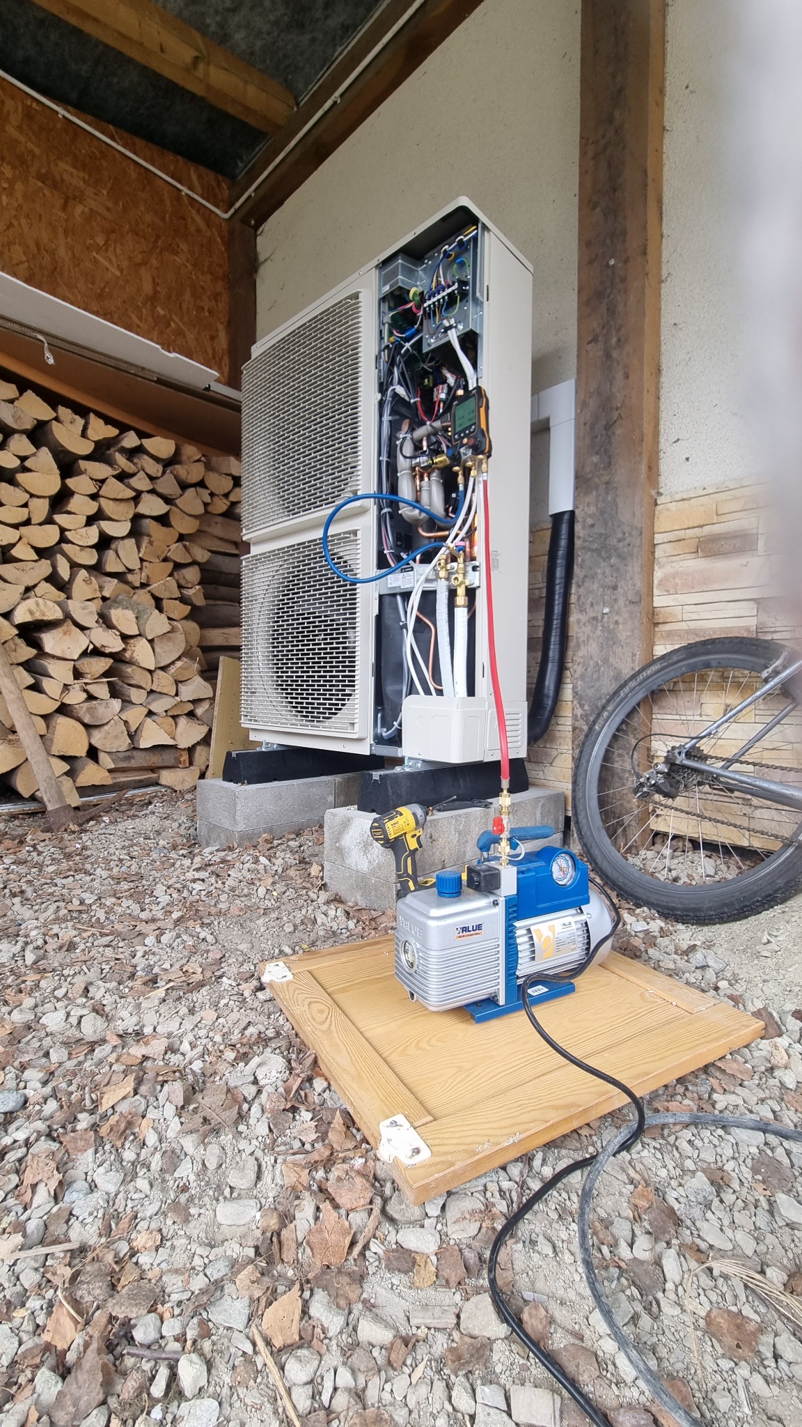 Instalacja zewnętrznej jednostki grzewczej z otwartą obudową, podłączona do pompy próżniowej VALUE na drewnianej płycie, w tle stos drewna i rower.