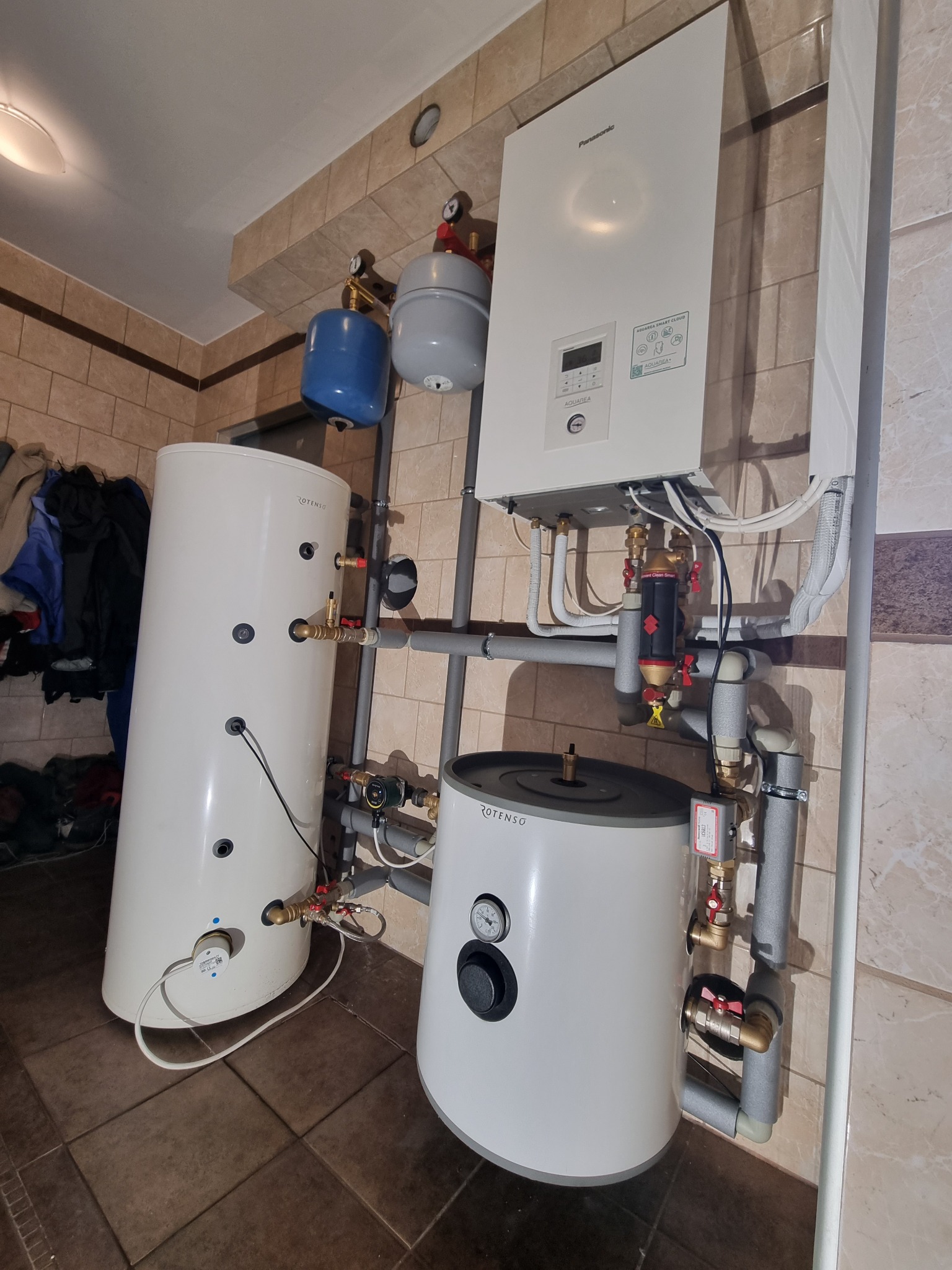 Zestaw urządzeń grzewczych w pomieszczeniu: biały zbiornik akumulacyjny Rotenso z czarnym manometrem, biały wiszący piec gazowy Panasonic Aquarea Smart Cloud, szare rury instalacyjne,...