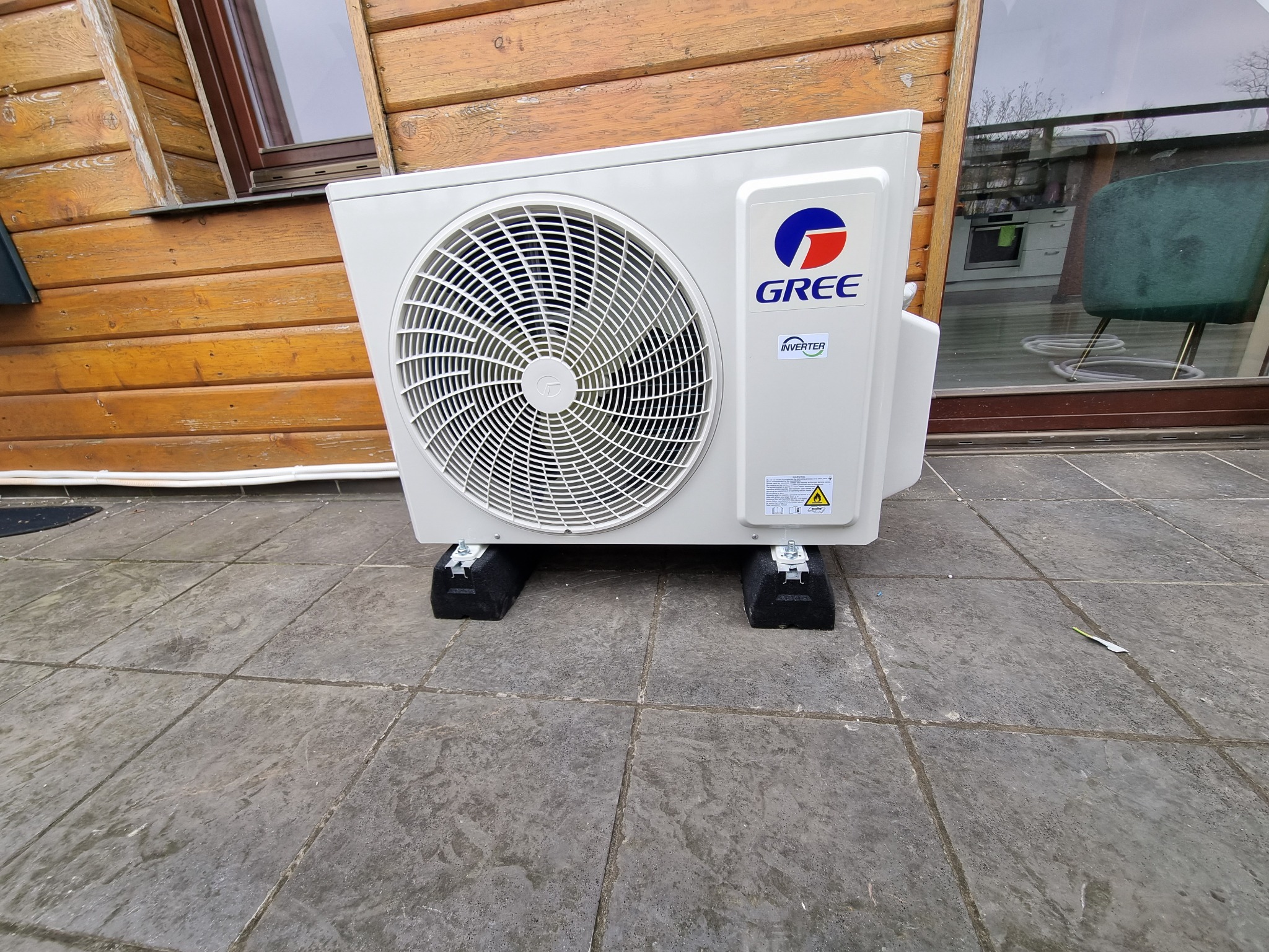 Zewnętrzna jednostka klimatyzacji marki Gree zamontowana na czarnych podkładkach, widoczna na tle drewnianej elewacji budynku i okna. W odbiciu szyby widoczny fragment wnętrza z krzesłem i kuchenną...