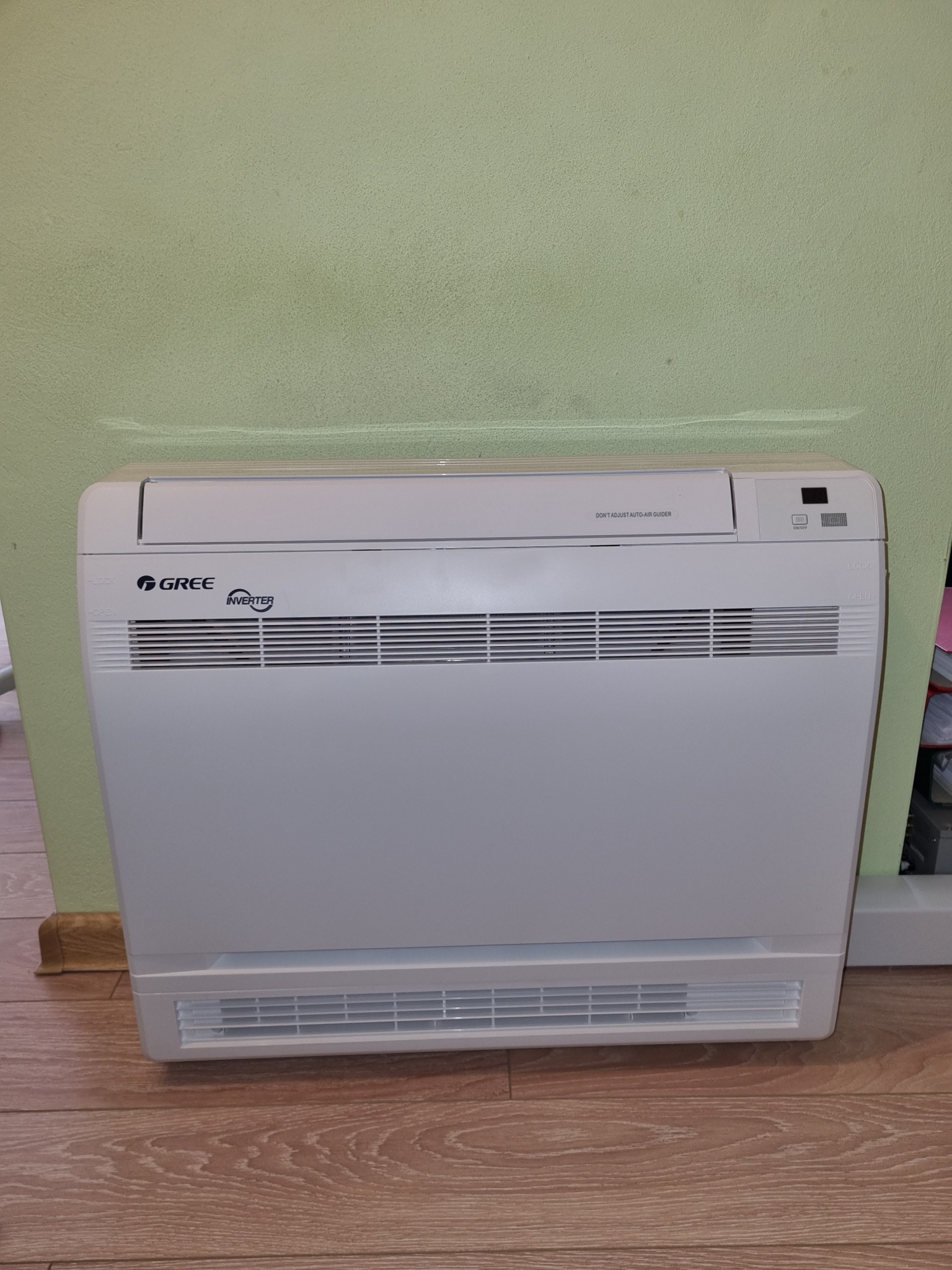 Biała, przypodłogowa jednostka klimatyzacyjna marki Gree Inverter, umieszczona przy zielonej ścianie i drewnianej podłodze.