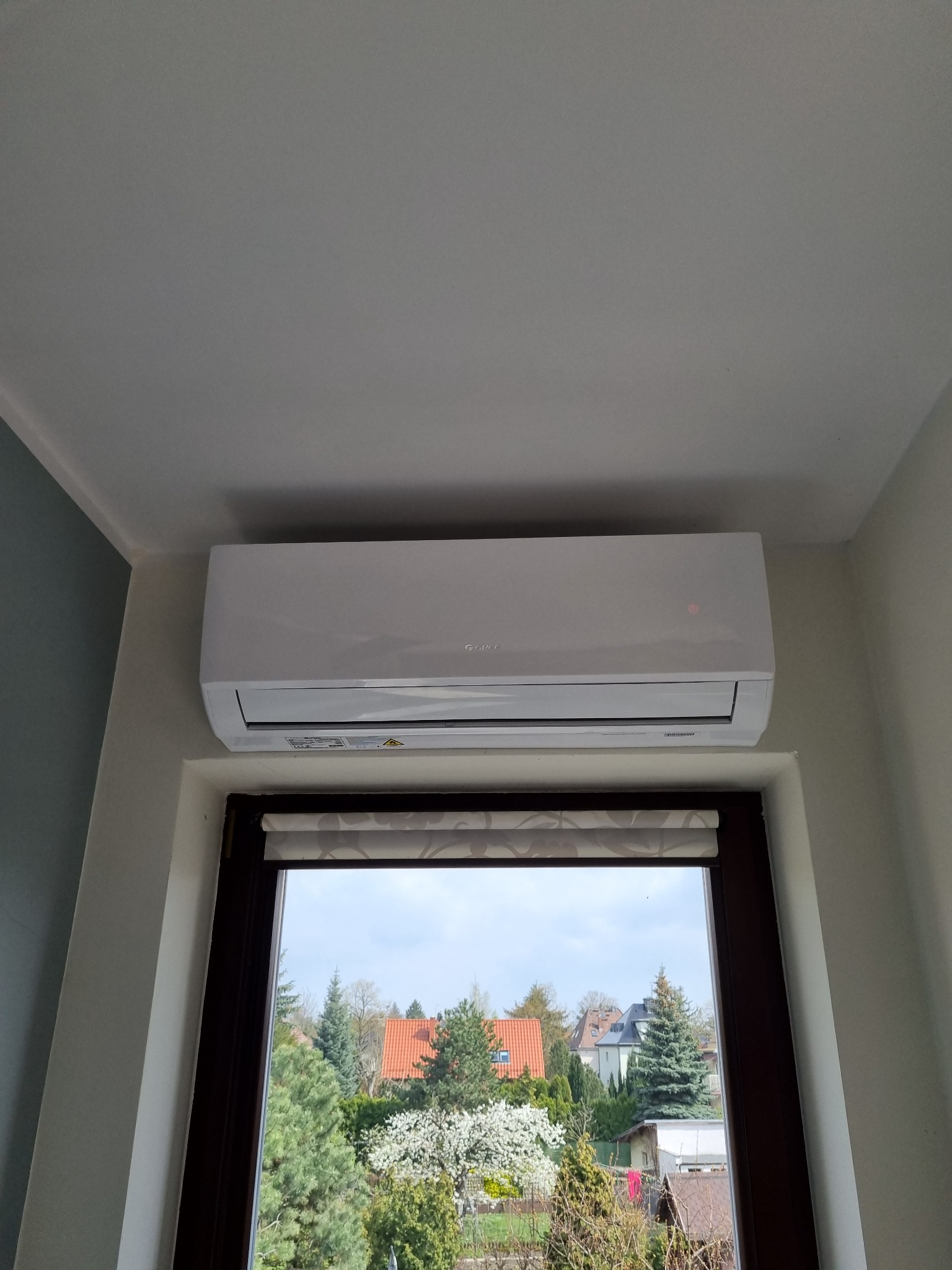Biały klimatyzator zamontowany nad oknem z widokiem na ogród i domy w tle. Urządzenie marki Gree z widoczną diodą kontrolną.