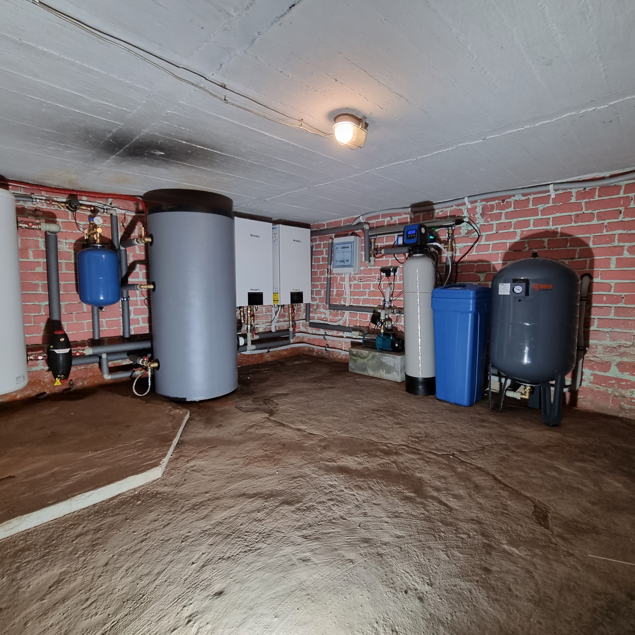 Instalacja urządzeń grzewczych i sanitarnych w piwnicy z ceglaną ścianą, widoczne zbiorniki, filtry, pompy, rury oraz dwa białe piece grzewcze.