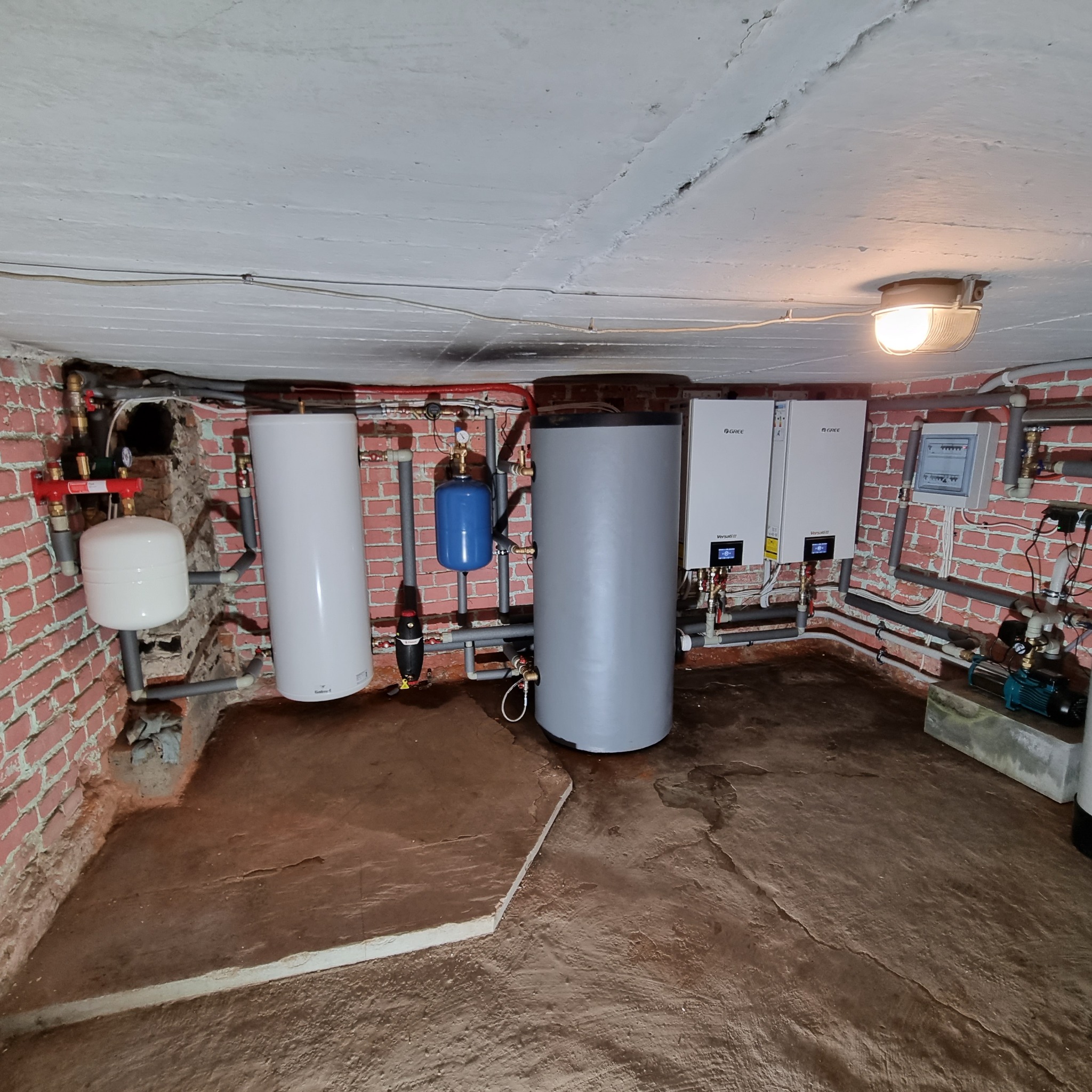 Zestaw urządzeń grzewczych w piwnicy z ceglanymi ścianami, obejmujący dwa białe piece Gree Versati III, szary zbiornik buforowy, biały zbiornik wyrównawczy, niebieski zbiornik ciśnieniowy...
