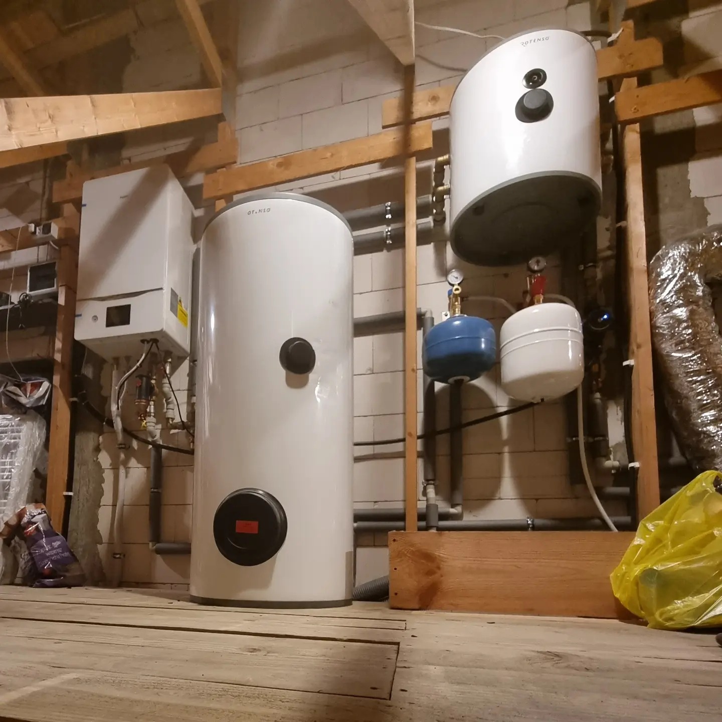 Instalacja kotła grzewczego i zasobników ciepłej wody użytkowej w kotłowni z drewnianą konstrukcją stropu i ścianami z bloczków. Widoczne elementy instalacji hydraulicznej i elektrycznej.