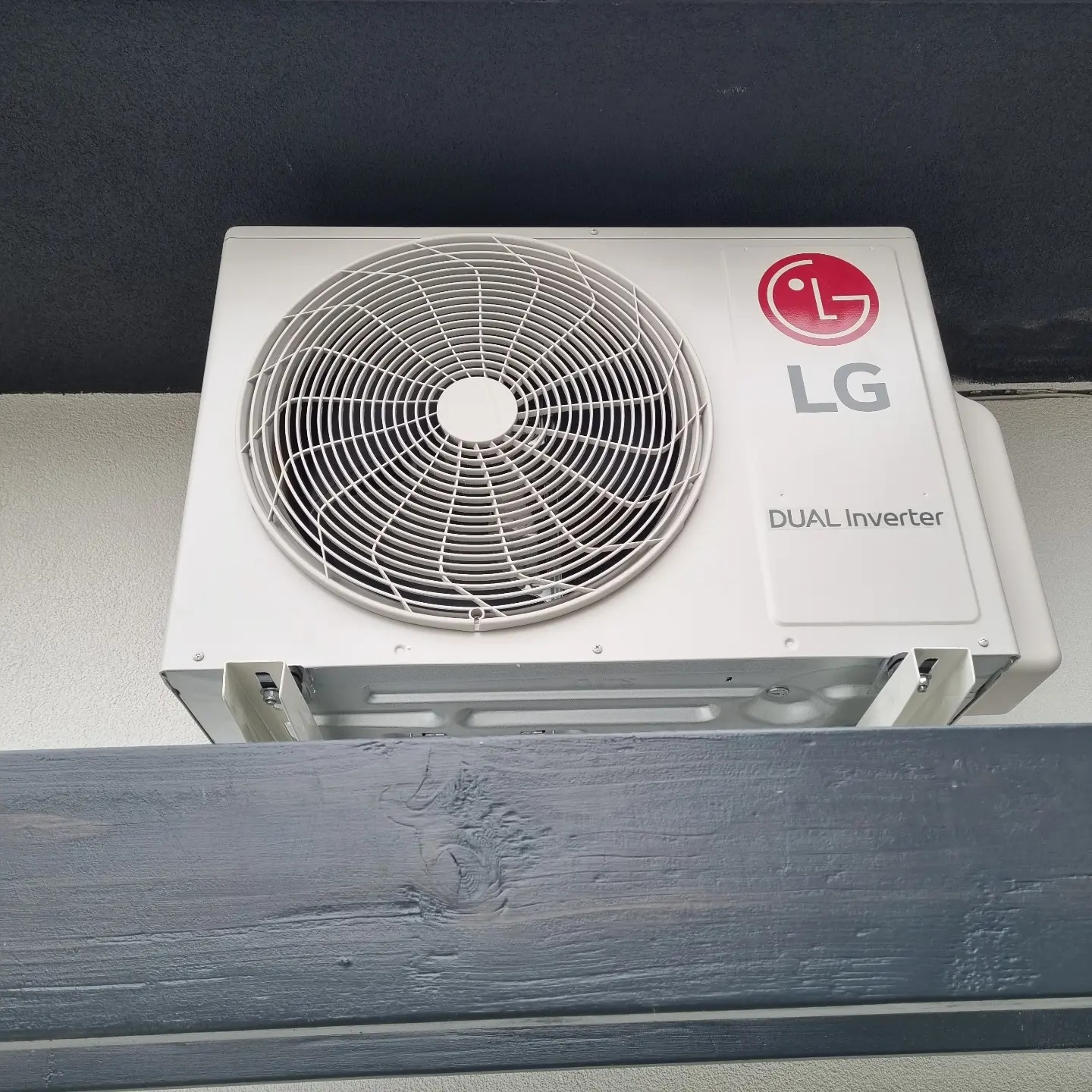 Biała jednostka zewnętrzna klimatyzacji LG Dual Inverter zamontowana na szarej, drewnianej belce pod ciemnym sufitem, widok z dołu.