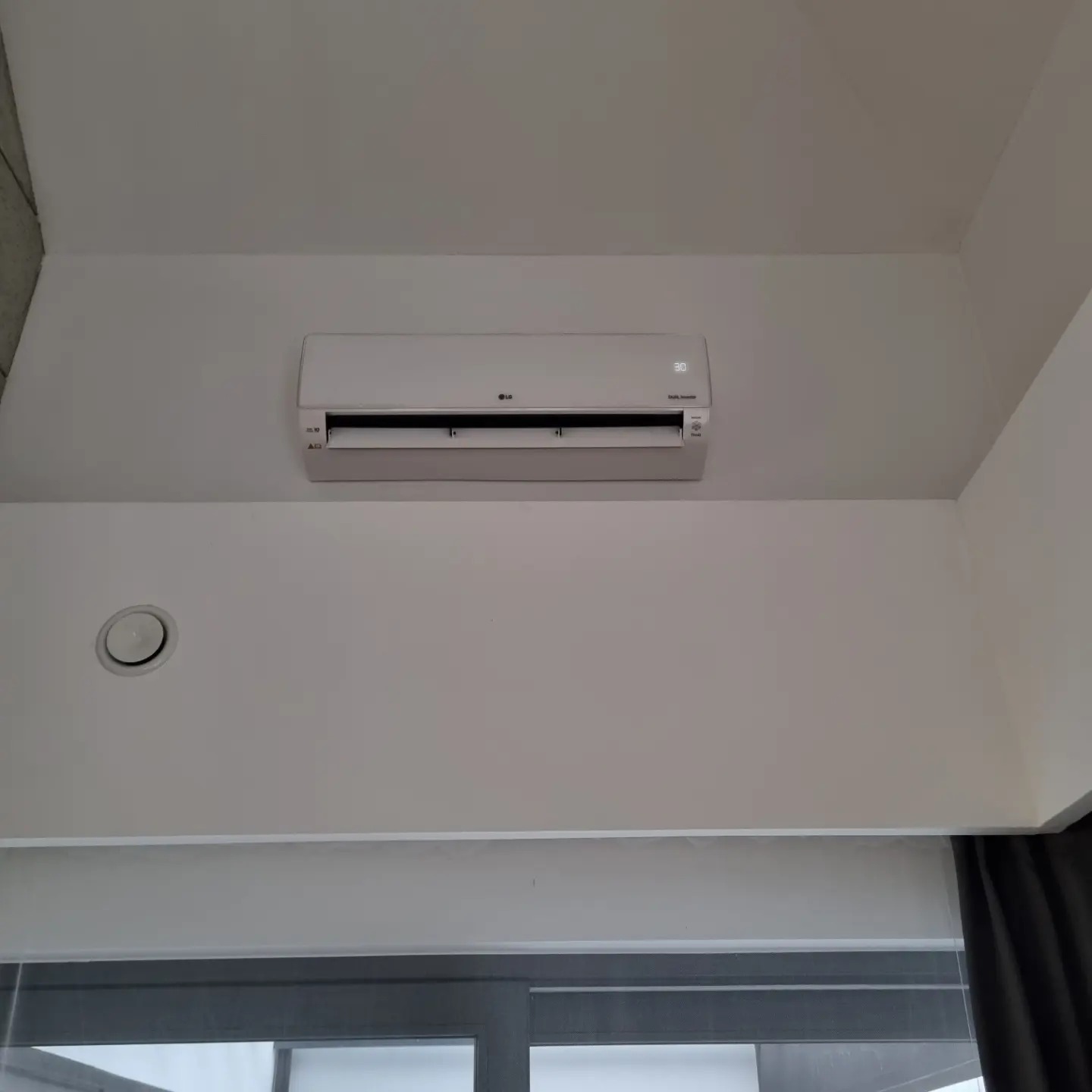 Biały klimatyzator LG Dual Inverter zamontowany pod sufitem w jasnym pomieszczeniu, widoczny fragment okna i zasłony.