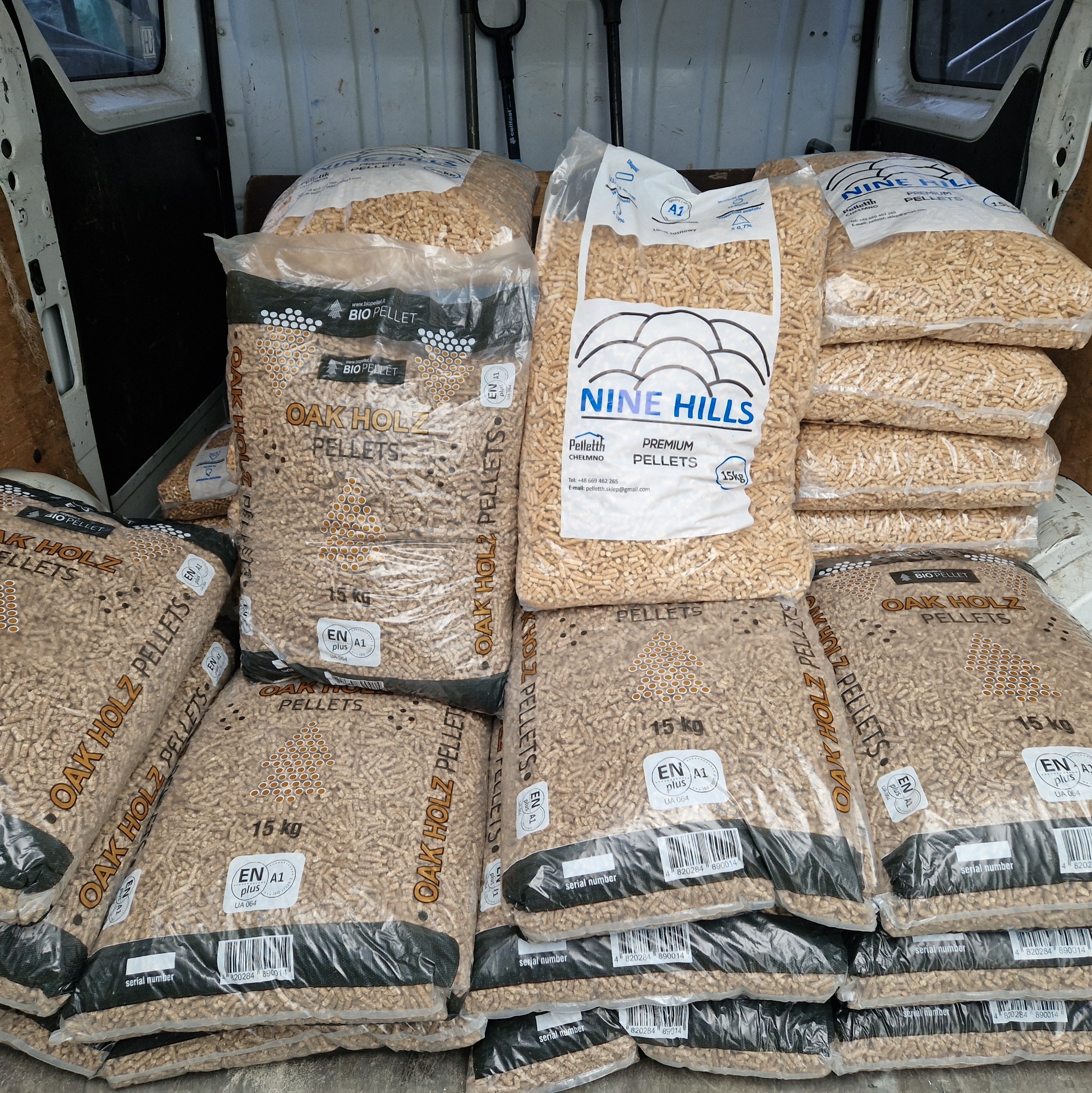 Wnętrze furgonetki wypełnione workami z pelletem drzewnym różnych marek, w tym Oak Holz i Nine Hills, z widocznymi oznaczeniami certyfikacji EN A1 Plus i wagą 15 kg na każdym worku.