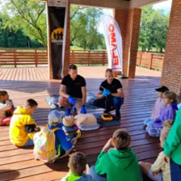 Instruktorzy w niebieskich rękawiczkach demonstrują użycie defibrylatora na manekinie podczas zajęć z pierwszej pomocy dla dzieci siedzących na drewnianej podłodze w zadaszonej przestrzeni.