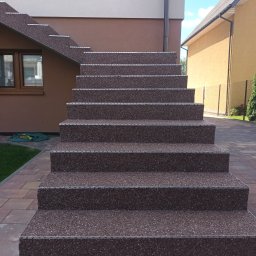 Zaczarowany kamyk - Zewnętrzne schody z brązowego kruszywa, wykończone metalowymi listwami, prowadzące do domu z beżowo-brązową elewacją, widok z dołu.