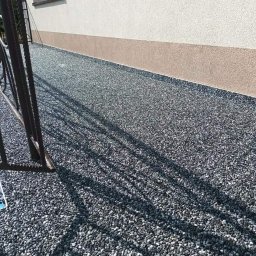 Zaczarowany kamyk - Wykończona powierzchnia z drobnych, szarych kamyczków przy ścianie budynku z widocznym fragmentem metalowej balustrady i cieniem drzewa.