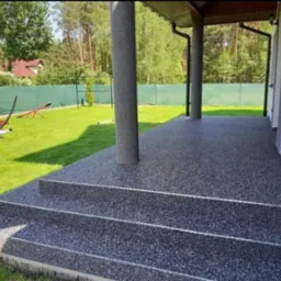 Szary, żywiczny taras z trzema stopniami, widok na trawnik z drewnianym placem zabaw i las w tle.