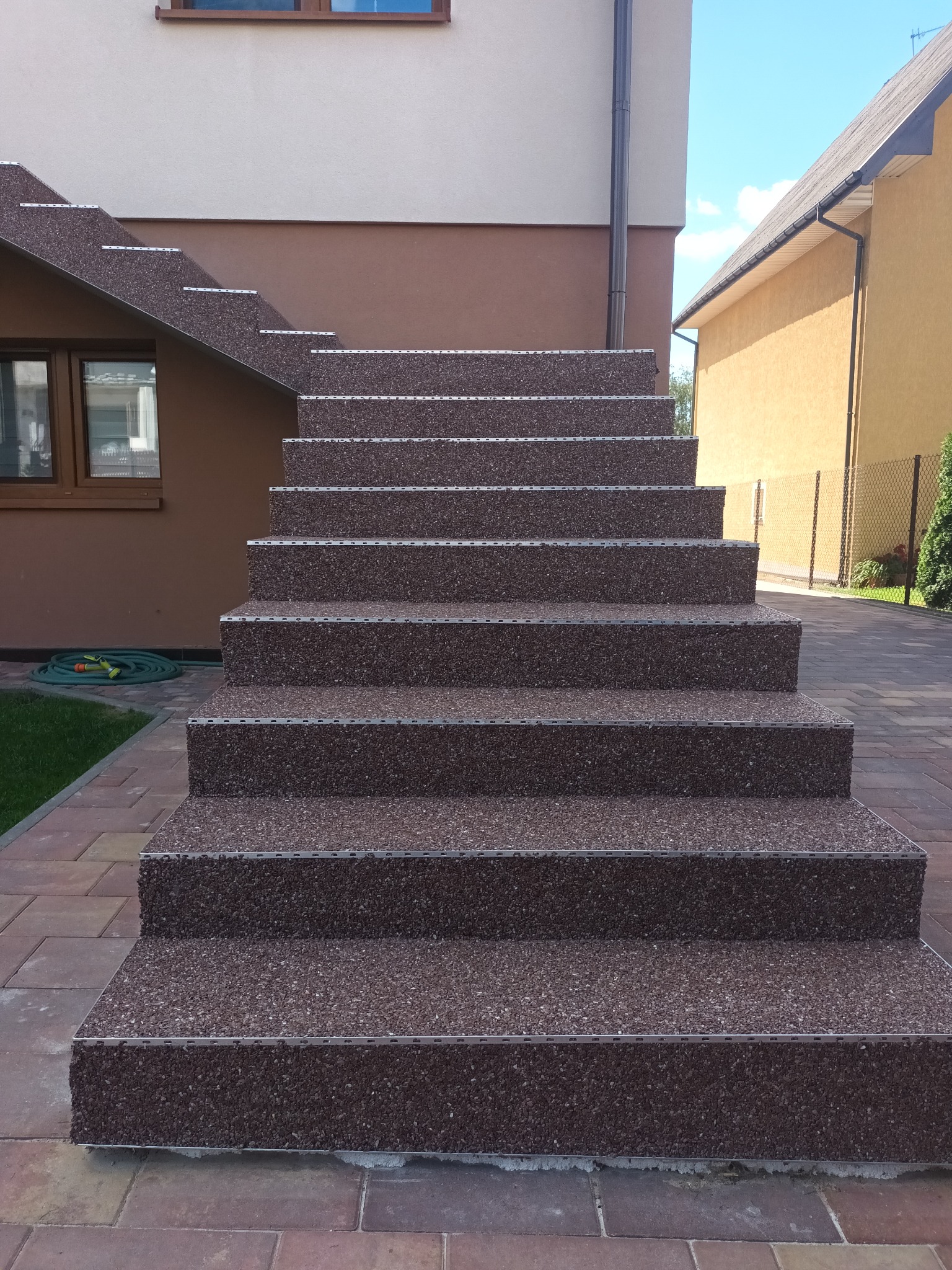 Zewnętrzne schody z brązowego kruszywa, wykończone metalowymi listwami, prowadzące do domu z beżowo-brązową elewacją, widok z dołu.