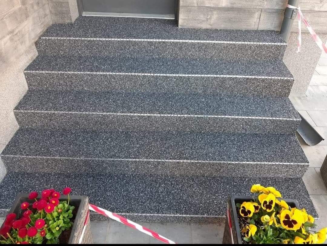 Zewnętrzne schody z szarego kamienia kompozytowego z metalowymi wykończeniami krawędzi stopnic, ozdobione donicami z czerwonymi i żółtymi bratkami.