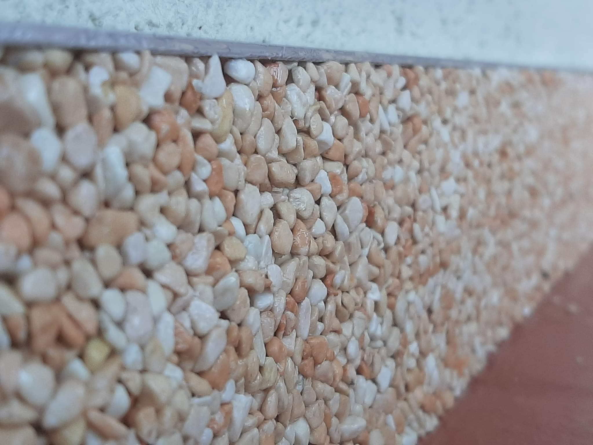 Dekoracyjna ściana wykończona mozaiką z drobnych, białych i beżowych kamyczków, zabezpieczona szarą listwą przy górnej krawędzi.