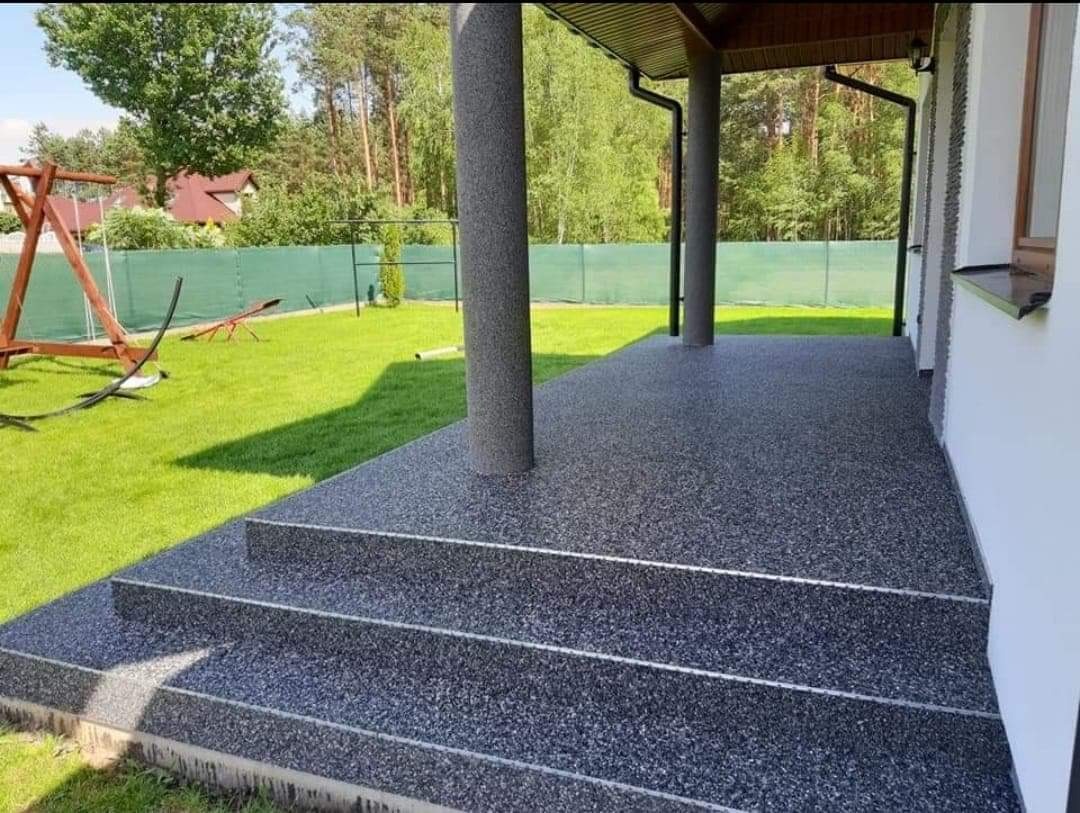 Szary, żywiczny taras z trzema stopniami, widok na trawnik z drewnianym placem zabaw i las w tle.