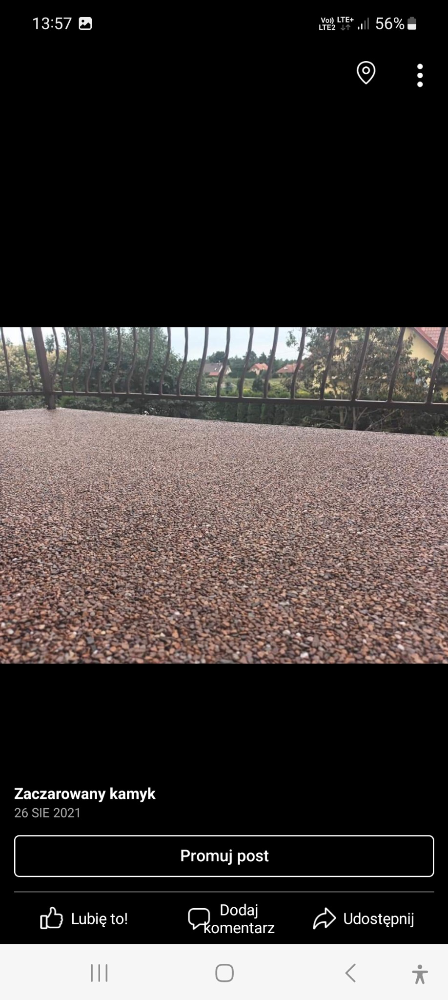 Wykończona powierzchnia tarasu z drobnych, brązowych kamyczków, widok zza metalowej balustrady, w tle zieleń drzew i zabudowania.