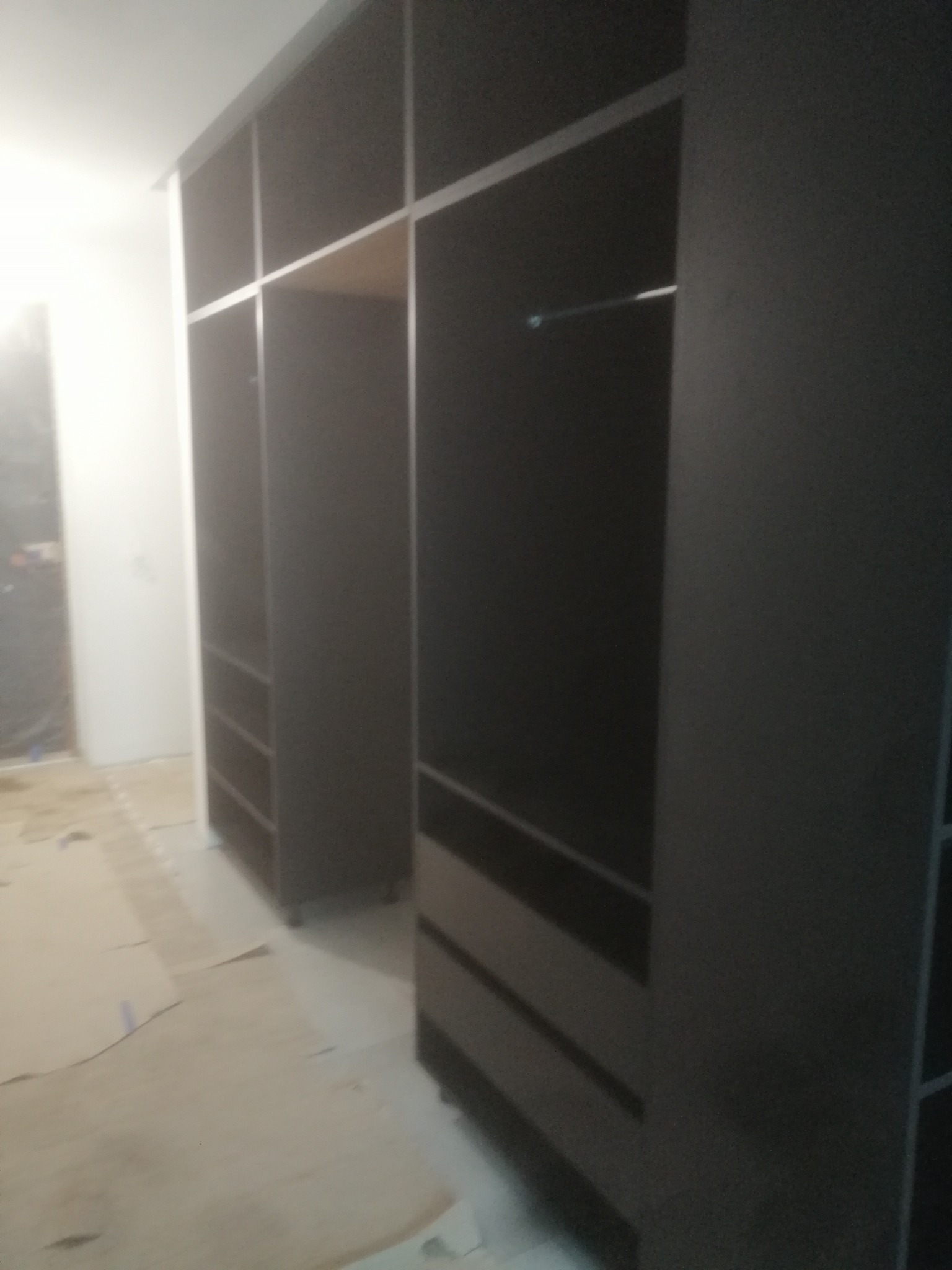 Ciemna, częściowo zamontowana szafa z szufladami i drążkiem na ubrania, widoczna na tle zabezpieczonej podłogi podczas remontu.