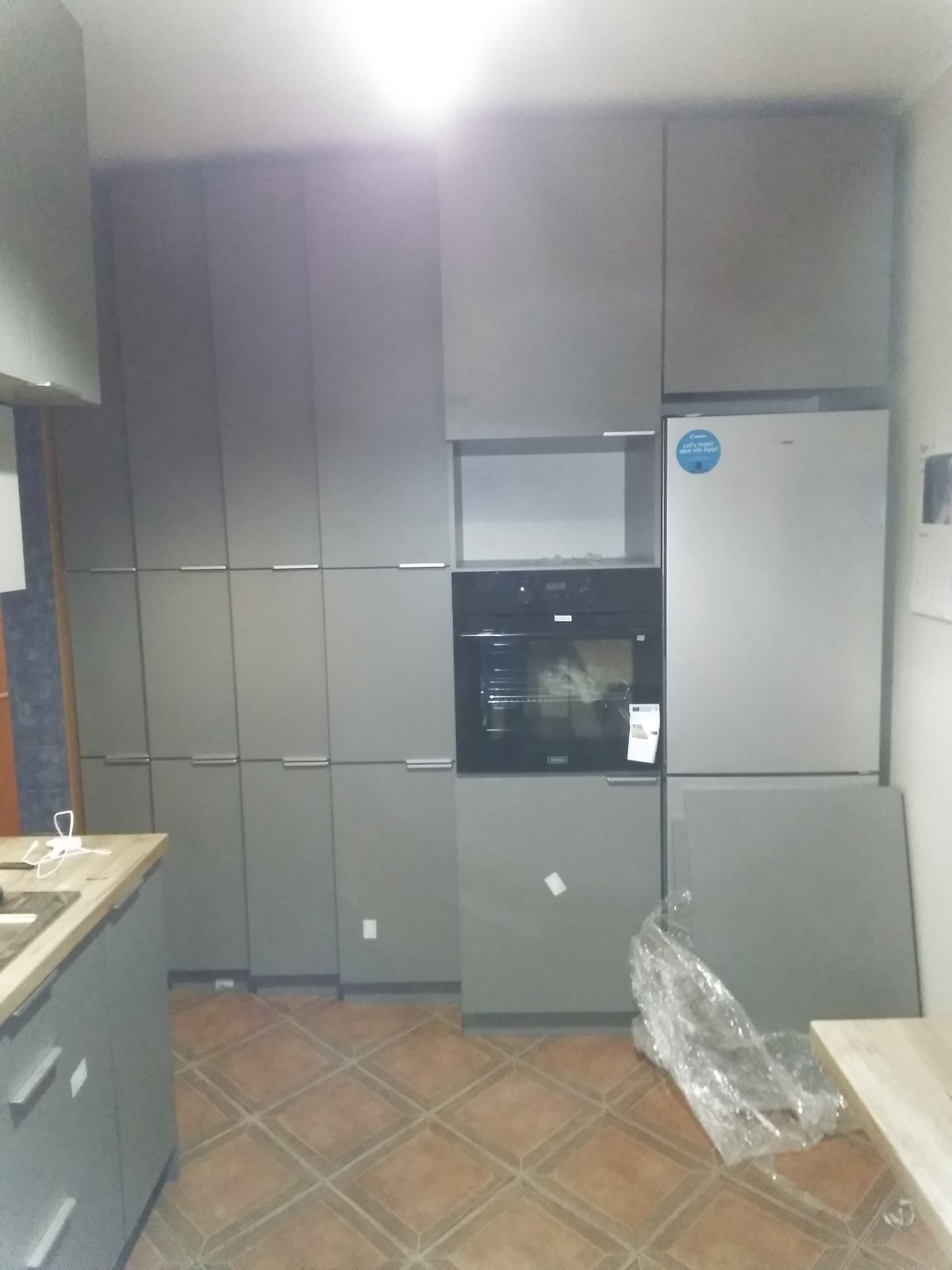 Szara zabudowa kuchenna z piekarnikiem i lodówką, widoczne płytki podłogowe w romby i elementy zabezpieczone folią