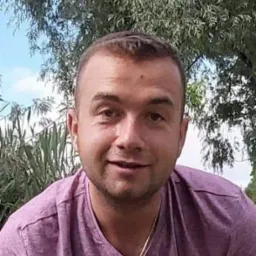 Portret uśmiechniętego mężczyzny w fioletowej koszulce na tle zieleni i błękitnego nieba.