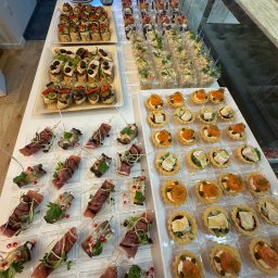 FAMILY - SERVICE YANA ANDRIUSHCHENKO - Bogaty wybór przekąsek cateringowych na białym stole: tartaletki z łososiem i brie, koreczki z szynką parmeńską i bakłażanem, sałatki warzywne w plastikowych kubeczkach oraz bruschetty z warzywami.