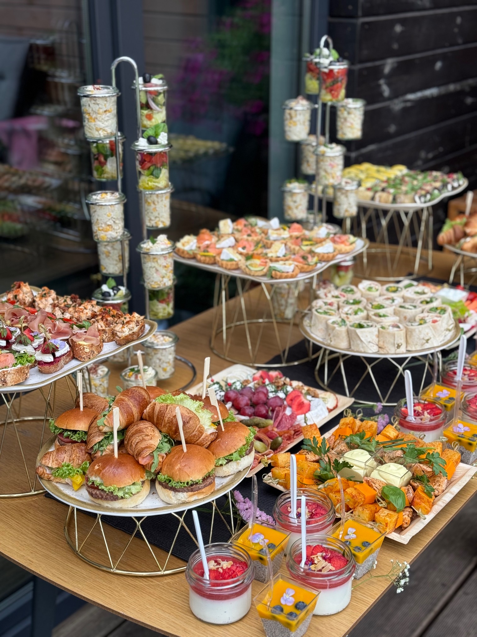Bogato zastawiony stół z różnorodnymi przekąskami: mini burgery z croissantami, tartaletki z łososiem, roladki z tortilli, sałatki w słoiczkach, desery w szklankach, koreczki z serem i winogronami,...