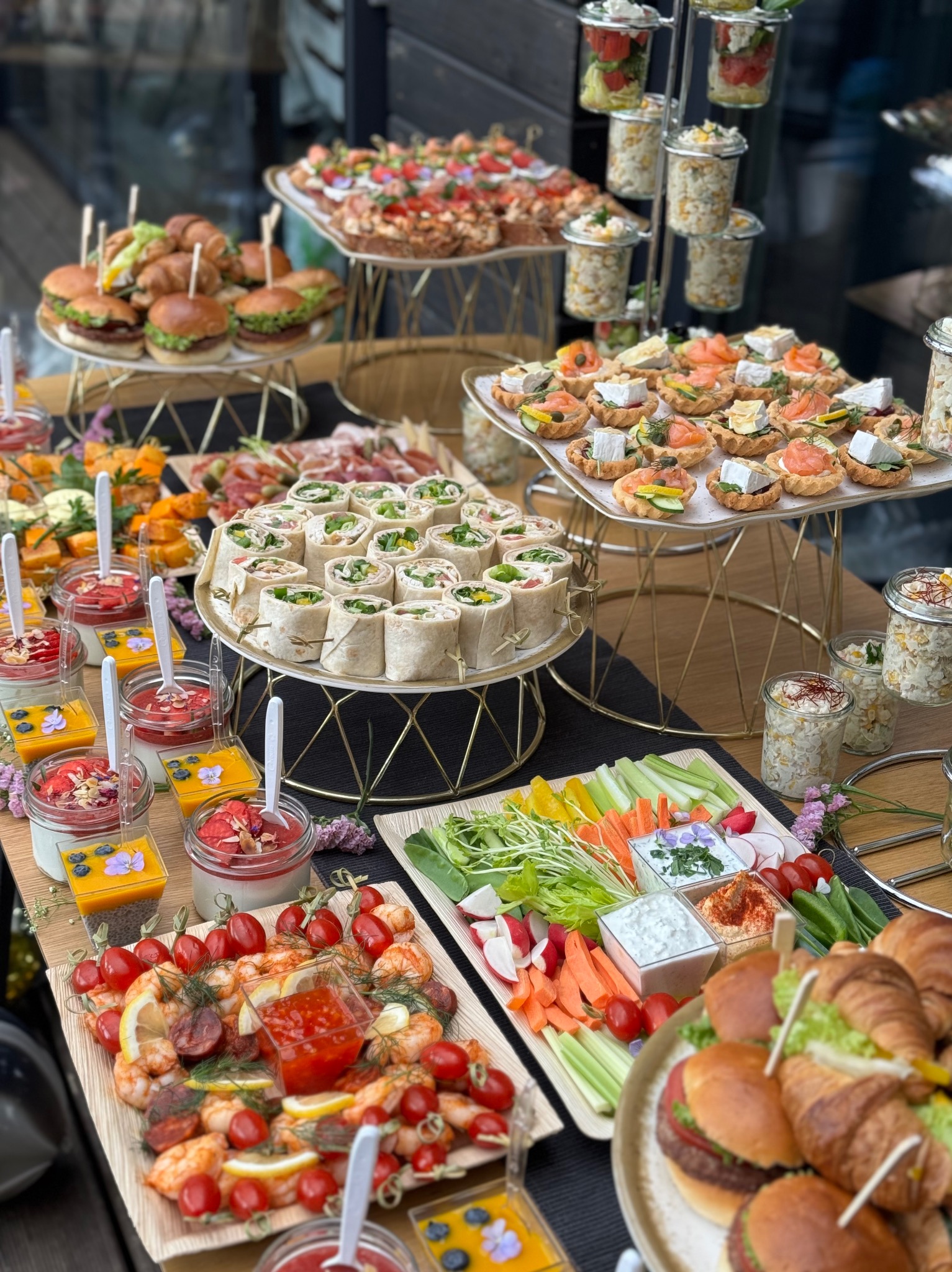Bogato zastawiony stół z różnorodnymi przekąskami: mini burgery, tortille, sałatki w słoiczkach, tartaletki z łososiem i camembertem, krewetki z pomidorami i dipami, oraz warzywa pokrojone w słupki...