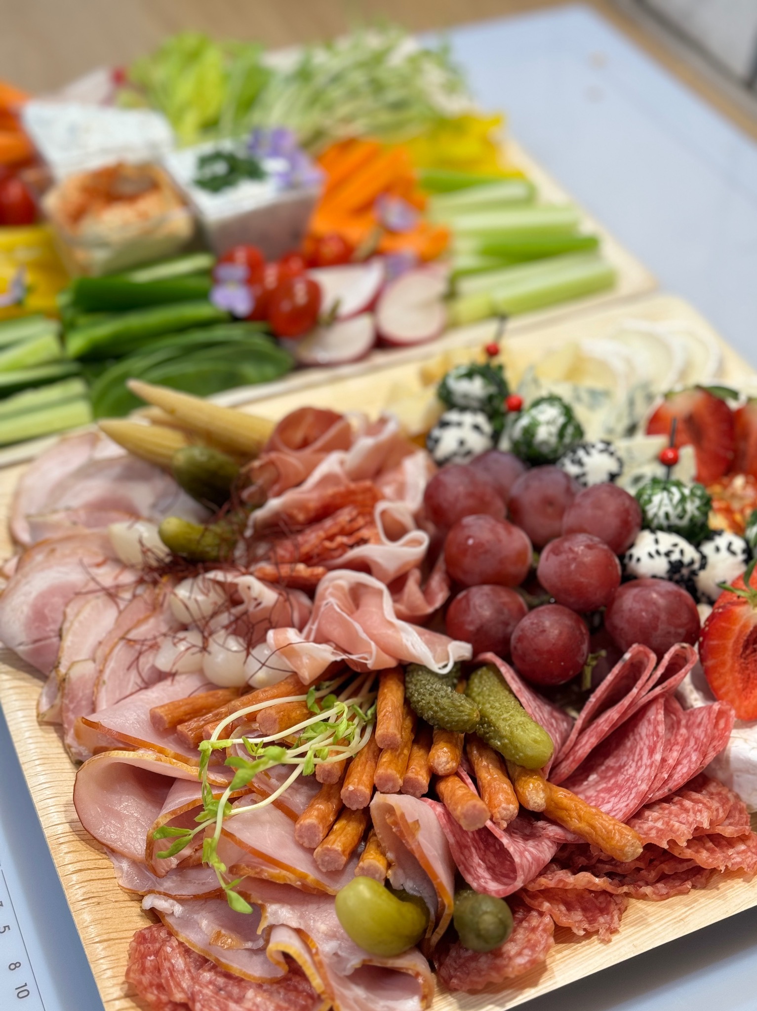 Dwie drewniane deski pełne różnorodnych przekąsek: warzywa (seler naciowy, marchewka, rzodkiewki, ogórki), dipy, szaszłyki z mini-mozzarelli, winogrona, truskawki oraz wędliny i sery, tworzące...