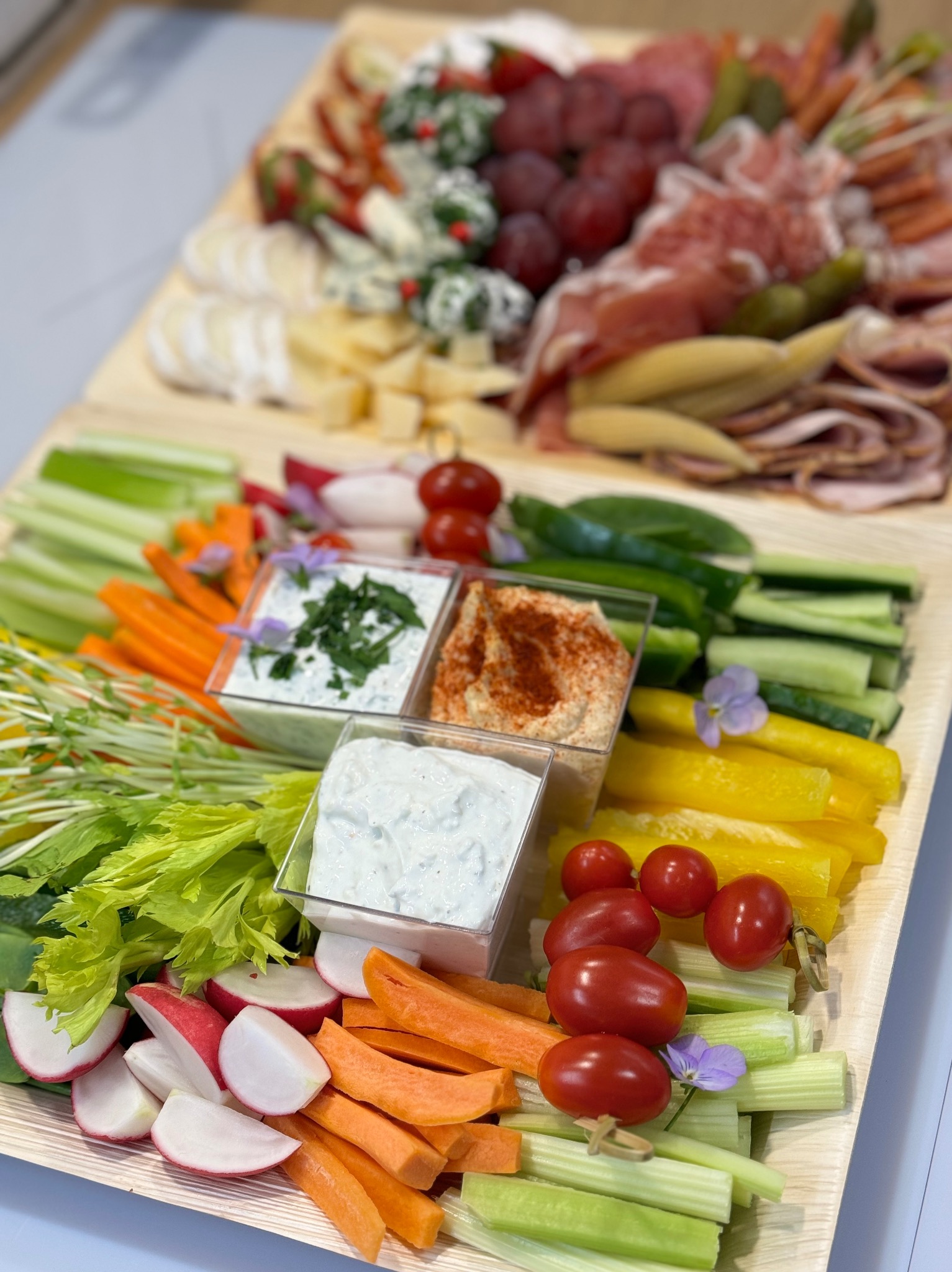 Elegancko ułożona drewniana deska z przekąskami: sery, wędliny, warzywa (seler naciowy, marchew, rzodkiewki, ogórki, papryka) i trzy sosy w małych pojemnikach, udekorowane drobnymi kwiatkami.