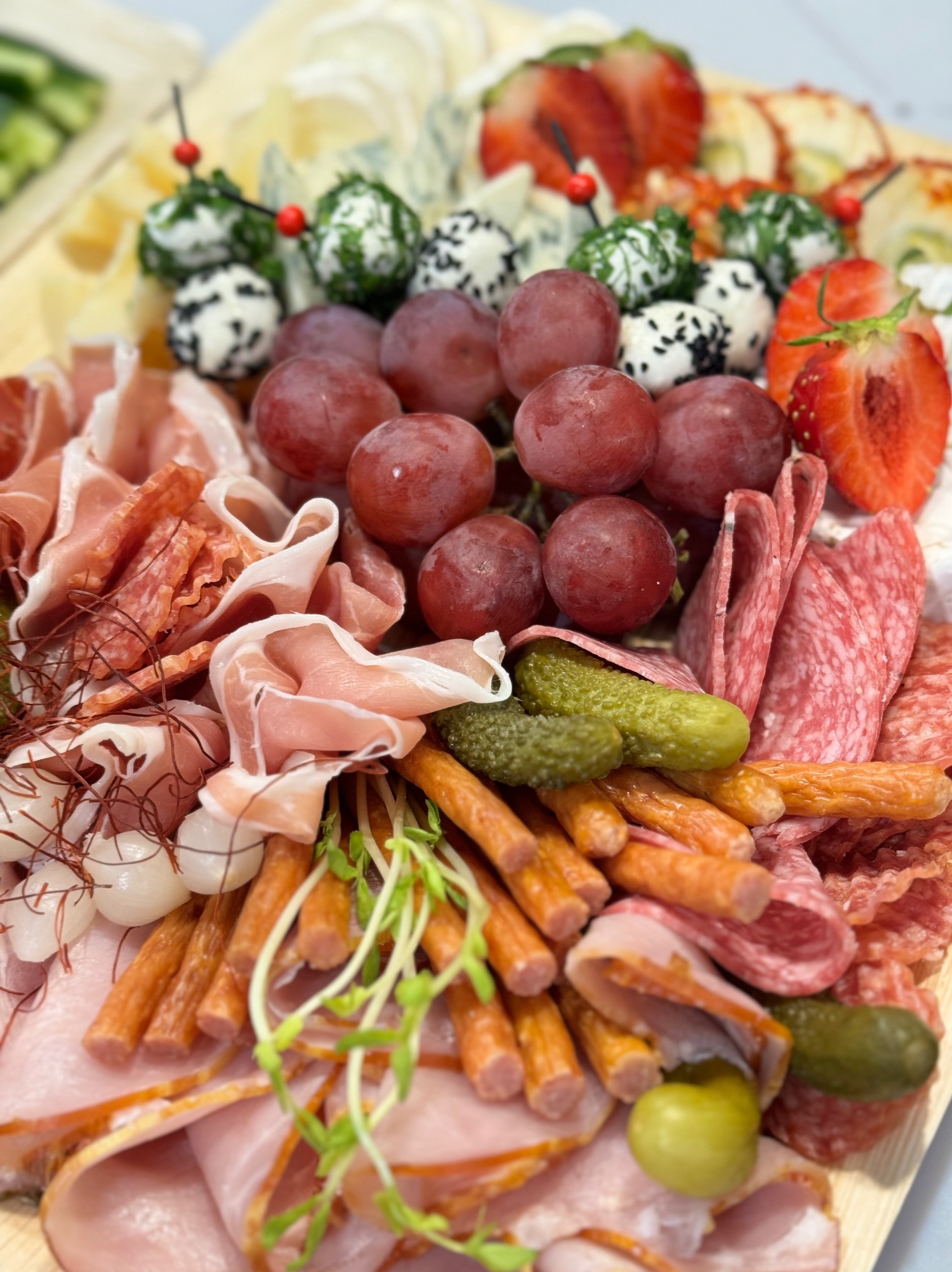 Bogato zaopatrzona deska serów, wędlin i owoców: kulki serowe z ziołami i czarnuszką, czerwone winogrona, plastry truskawek, szynka parmeńska, salami, kabanosy, pikle, cebulki marynowane i świeże...