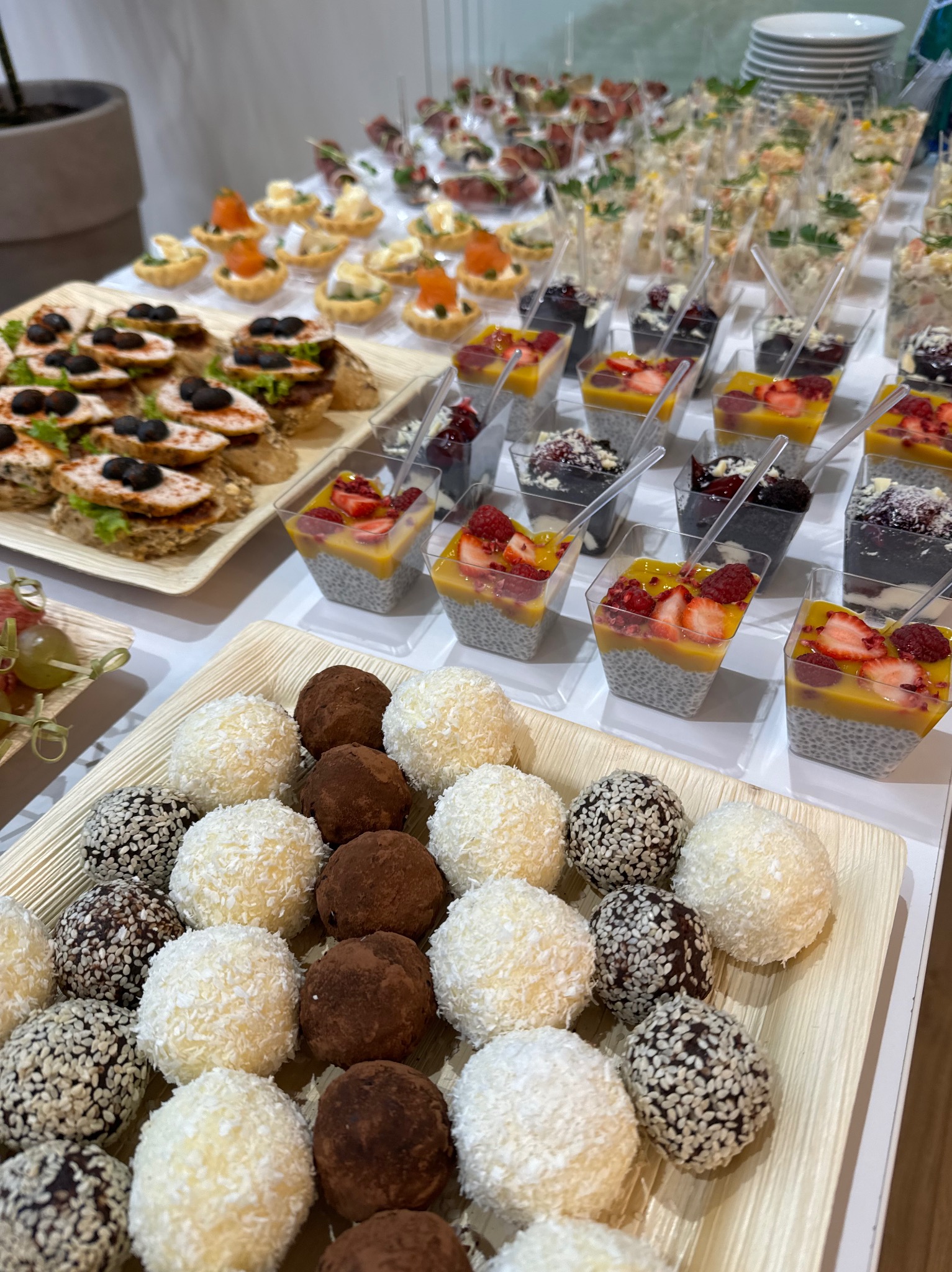 Różnorodne przekąski na przyjęciu: kanapki z oliwkami, mini tartaletki, sałatki w kubeczkach, desery chia z owocami i trufle kokosowe, sezamowe i kakaowe na drewnianych tackach.