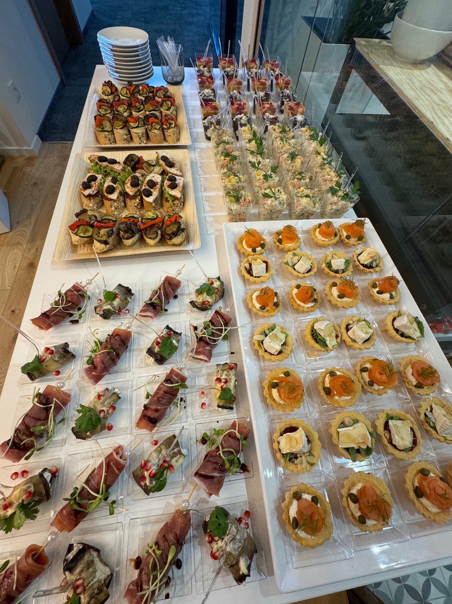 Bogaty wybór przekąsek cateringowych na białym stole: tartaletki z łososiem i brie, koreczki z szynką parmeńską i bakłażanem, sałatki warzywne w plastikowych kubeczkach oraz bruschetty z warzywami.