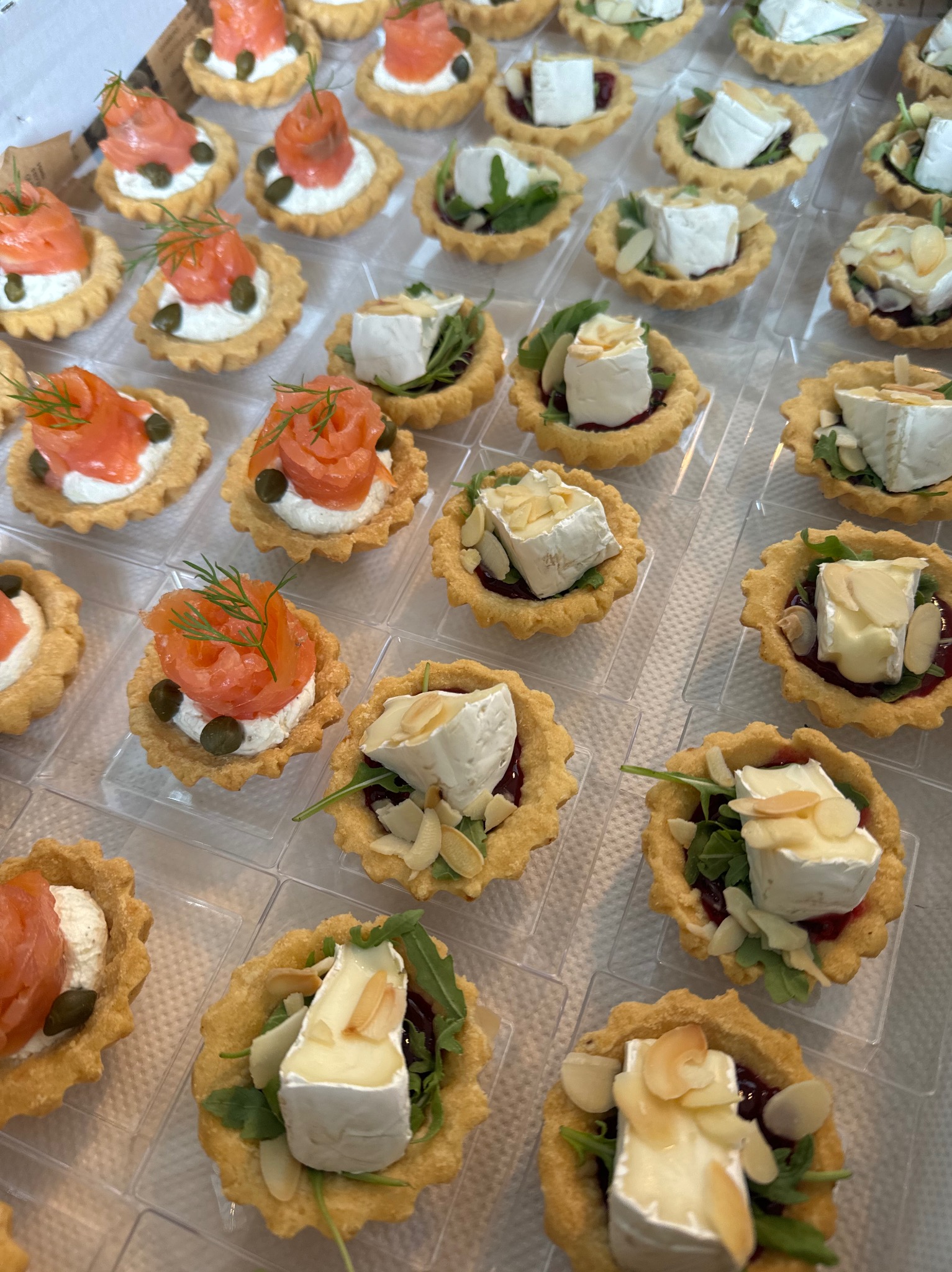 Mini tartaletki z łososiem, kaparami i koperkiem oraz z serem brie, rukolą, dżemem i migdałami, ułożone w przezroczystych przegródkach.
