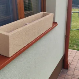 Beżowa donica na parapecie okna z brązową ramą, widoczna elewacja budynku o jasnej, ziarnistej strukturze i brązowa rynna. W tle trawnik i trampolina.