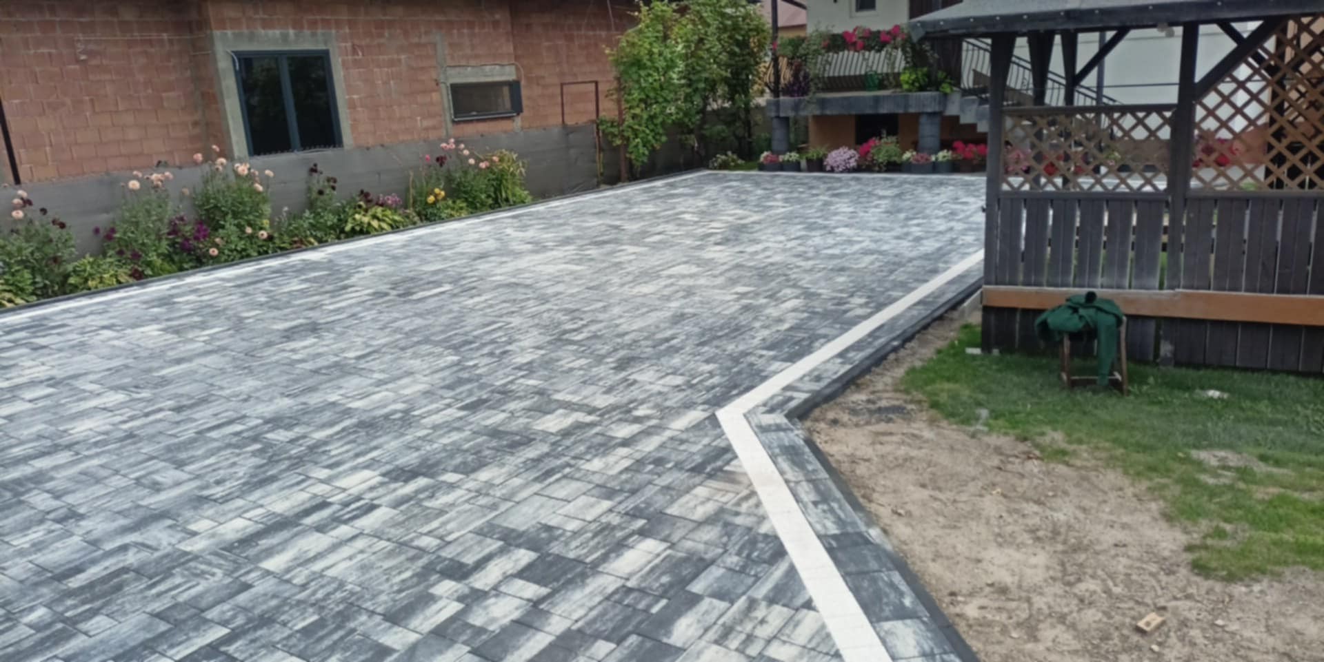 Ułożona kostka brukowa w szaro-białej tonacji, z białym obrzeżem, prowadząca do altany. Po bokach rabaty kwiatowe i budynek z cegły.