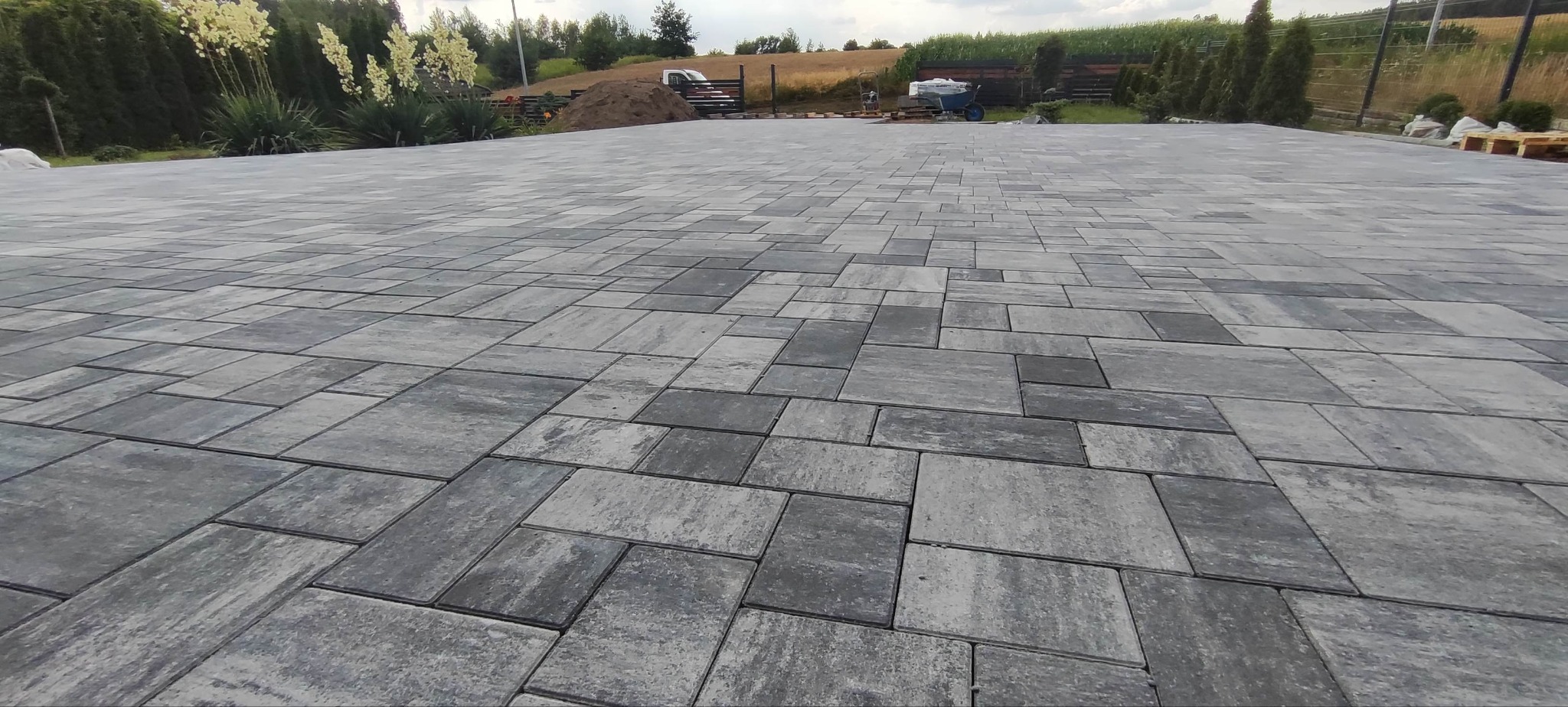 Ułożona, szara kostka brukowa na podjeździe, z widocznym tłem zieleni i elementami krajobrazu. Różne rozmiary kostek tworzą wzór.