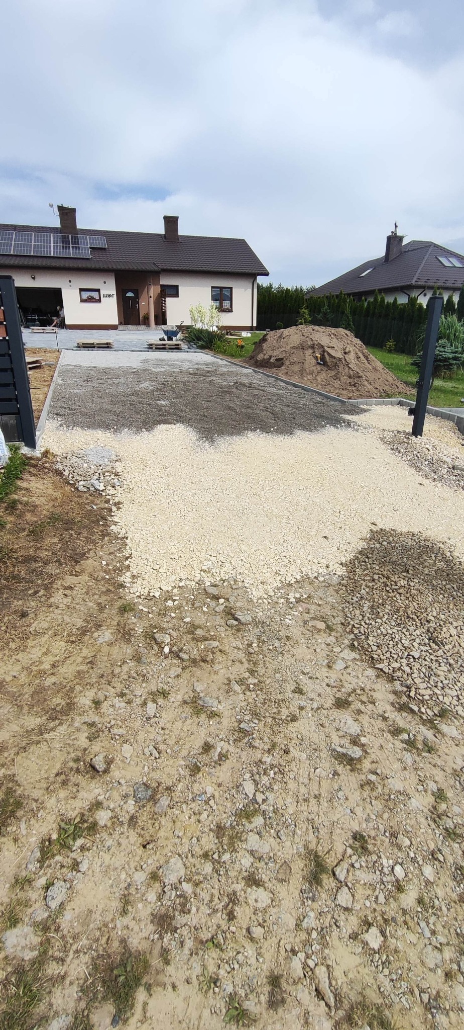 Etap przygotowania podłoża pod kostkę brukową: wysypany jasny żwir i ciemniejszy grys. W tle dom z panelami słonecznymi. Widoczny fragment ułożonej kostki.