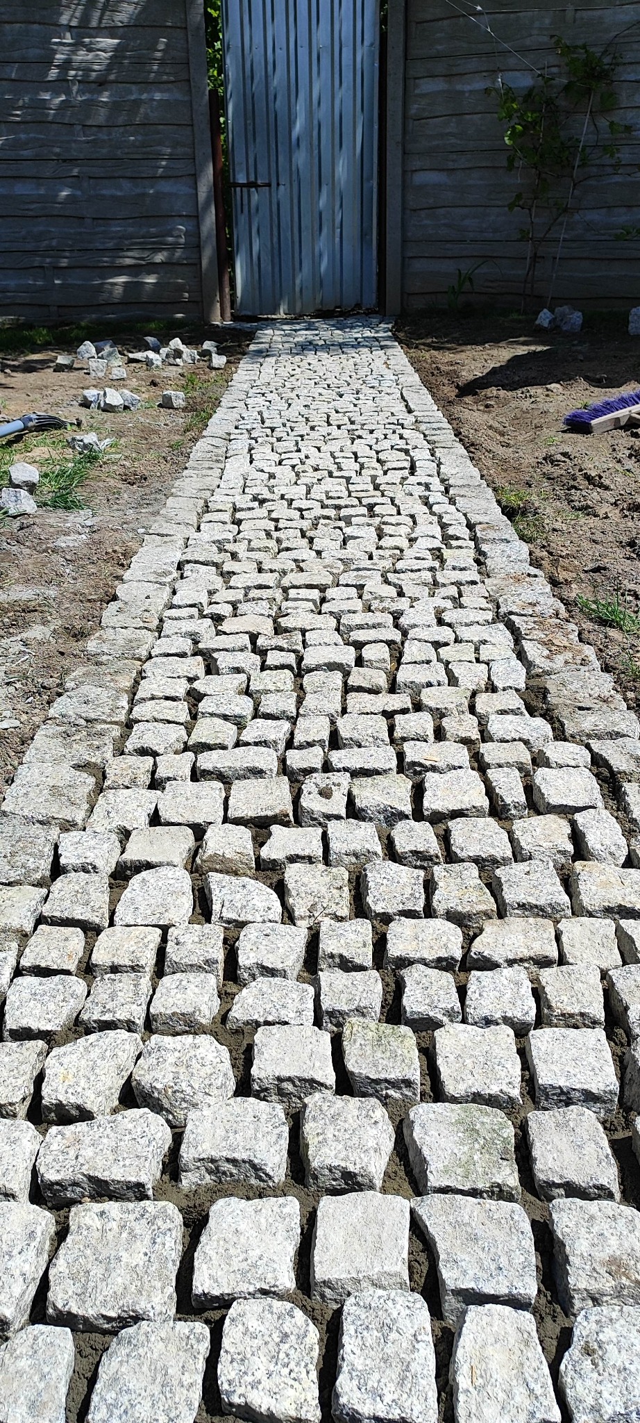Ścieżka z jasnoszarej kostki granitowej prowadząca do blaszanej bramy, widok z perspektywy osoby stojącej na początku ścieżki, obok leżą pojedyncze kostki.