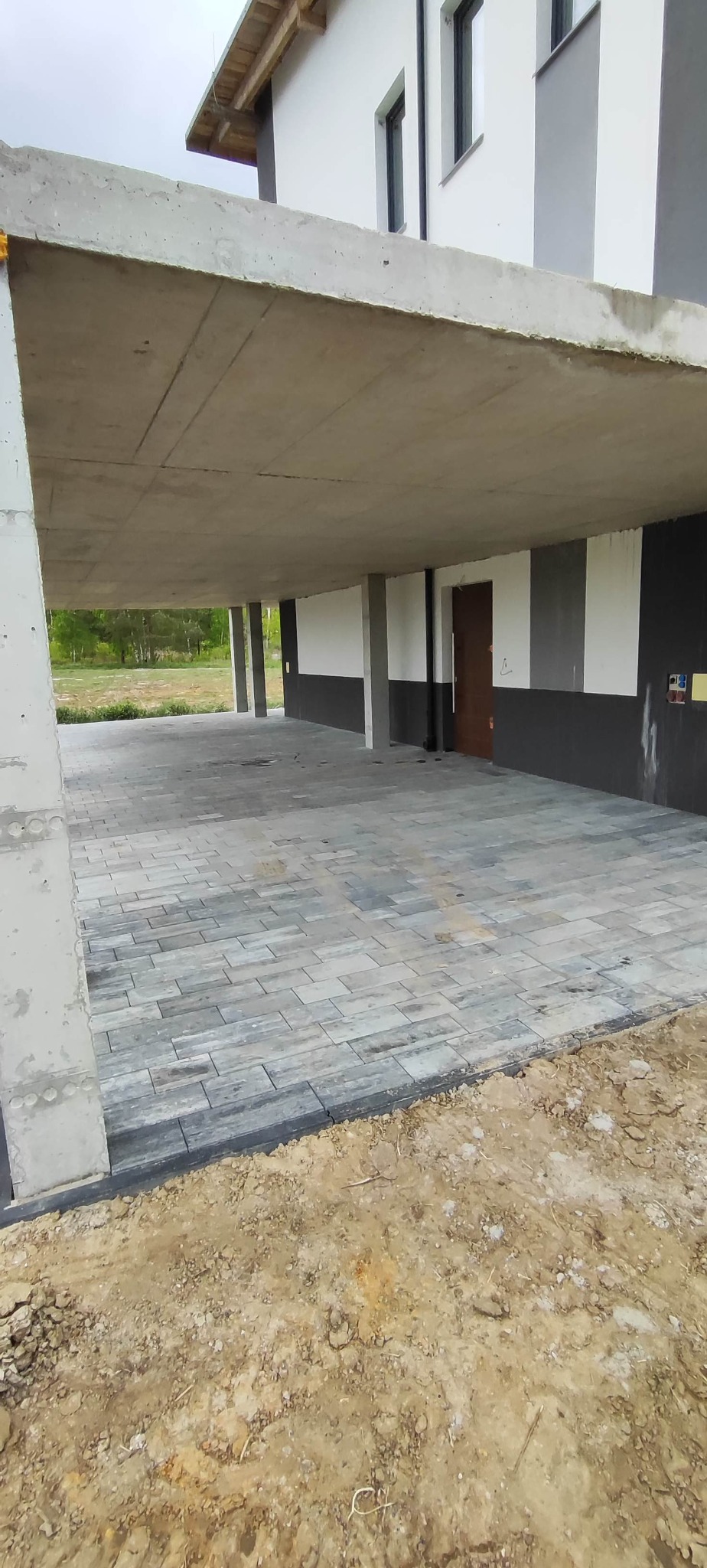 Ułożona kostka brukowa w szaro-niebieskiej tonacji pod betonowym zadaszeniem, widok z dołu, świeżo po realizacji, z pozostawioną ziemią wokół.