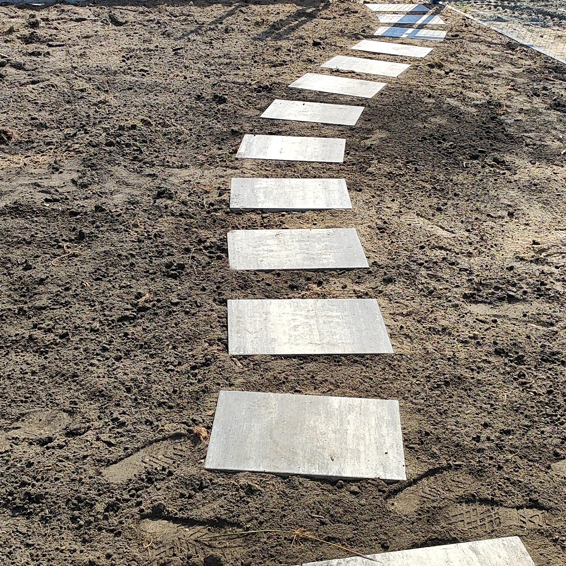 Ścieżka z szarych płyt ułożona na ziemi, tworząca chodnik w ogrodzie. Płyty prostokątne, ułożone w regularnych odstępach, prowadzą w głąb kadru. Widoczne ślady butów na ziemi.