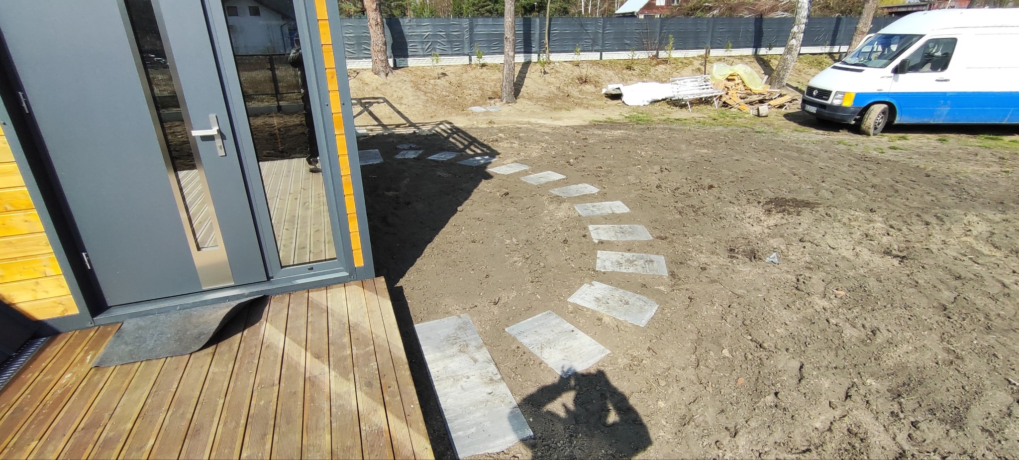Ścieżka z płyt kamiennych prowadząca od drewnianego tarasu do ogrodu. W tle widoczny samochód dostawczy i ogrodzenie z siatki.