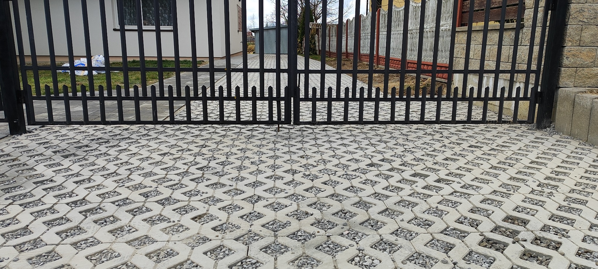 Szara ażurowa kostka brukowa wypełniona drobnym kamieniem, widoczna przez czarną, metalową bramę wjazdową. W tle budynki i zieleń. Kąt ujęcia z dołu.