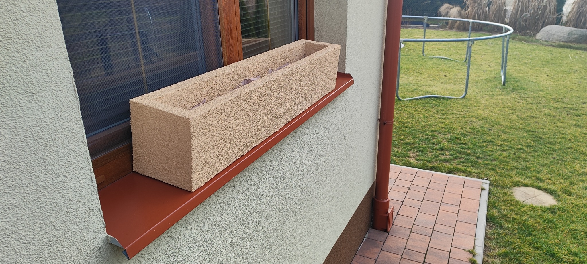 Beżowa donica na parapecie okna z brązową ramą, widoczna elewacja budynku o jasnej, ziarnistej strukturze i brązowa rynna. W tle trawnik i trampolina.