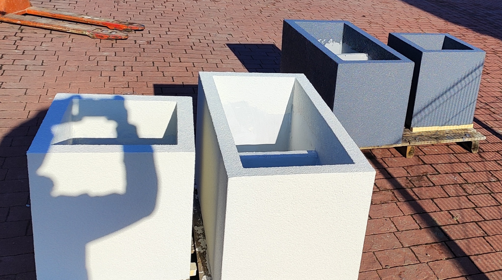 Cztery betonowe donice (dwie białe, dwie szare) na kostce brukowej, ustawione na palecie i widłach wózka widłowego, w jasnym oświetleniu słonecznym.