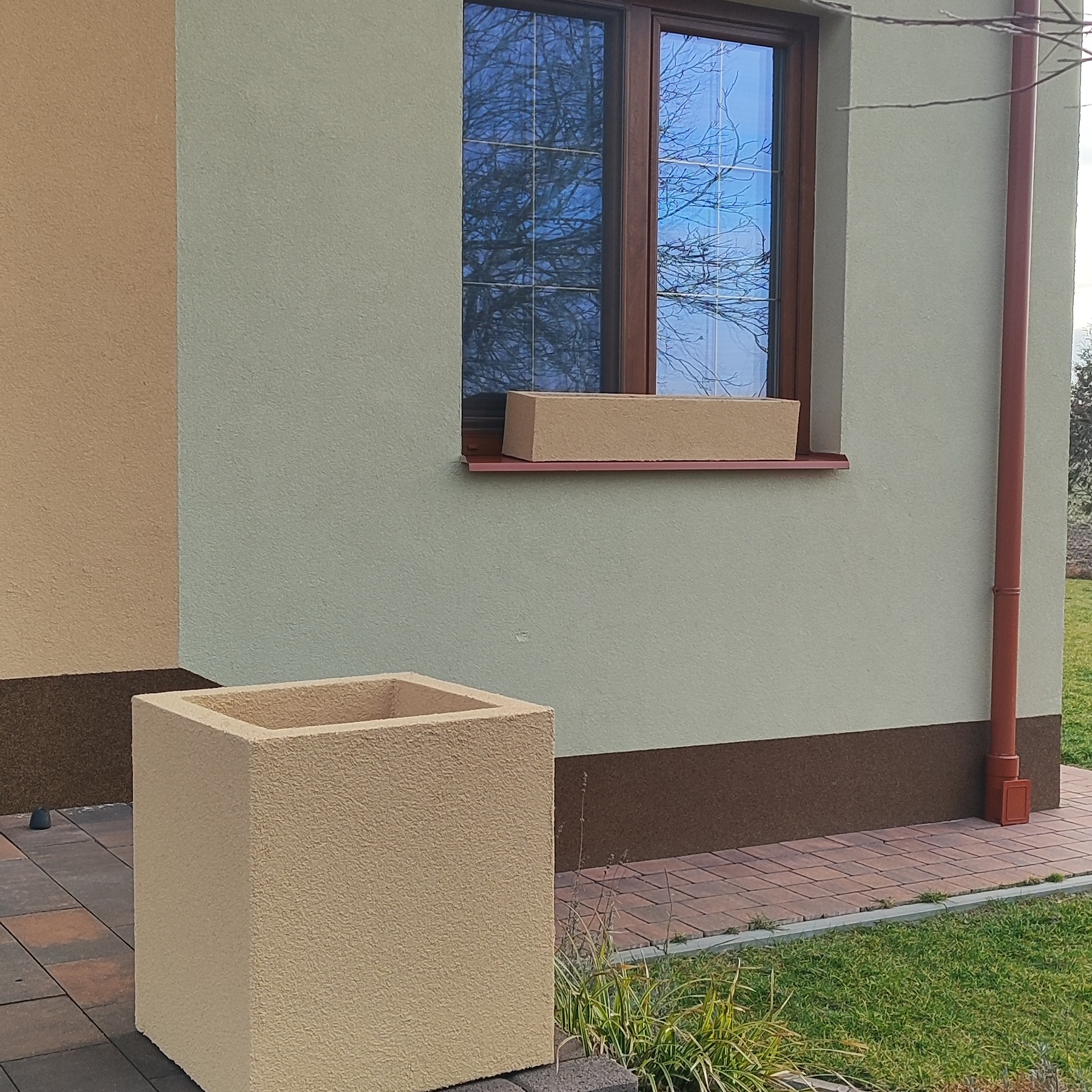 Ocieplona elewacja domu w odcieniach beżu i zieleni, z widoczną donicą na parapecie i kwadratową donicą na kostce brukowej. Widoczny fragment rynny.