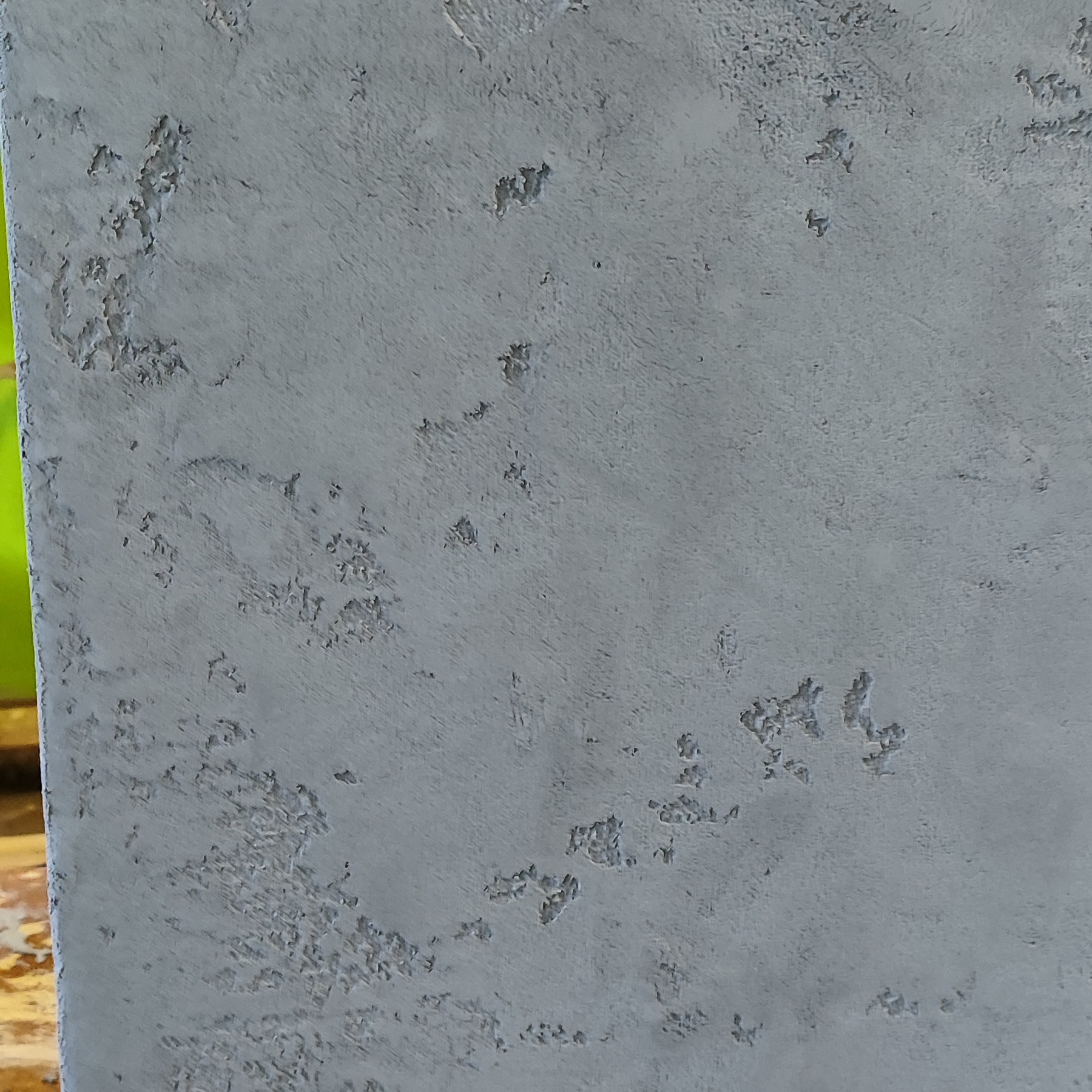 Szara, chropowata faktura ściany z bliska. Delikatne nierówności i cieniowania tworzą unikalny wzór. Widoczny fragment zieleni w tle.