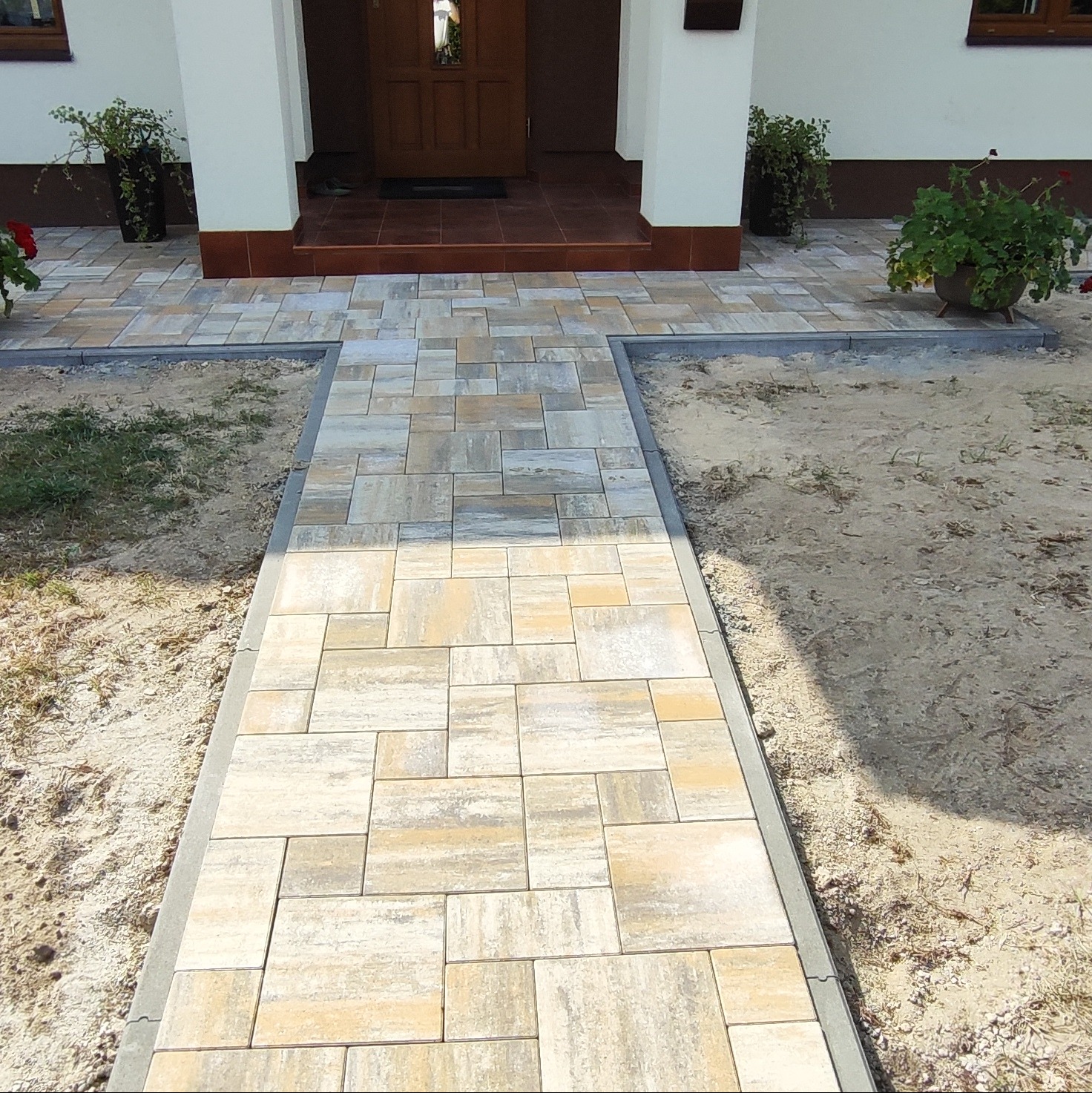 Ścieżka z kostki brukowej prowadzi do drzwi wejściowych. Jasne, prostokątne kostki ułożone w regularny wzór. Obrzeża z szarej kostki. Donice z roślinami po bokach.